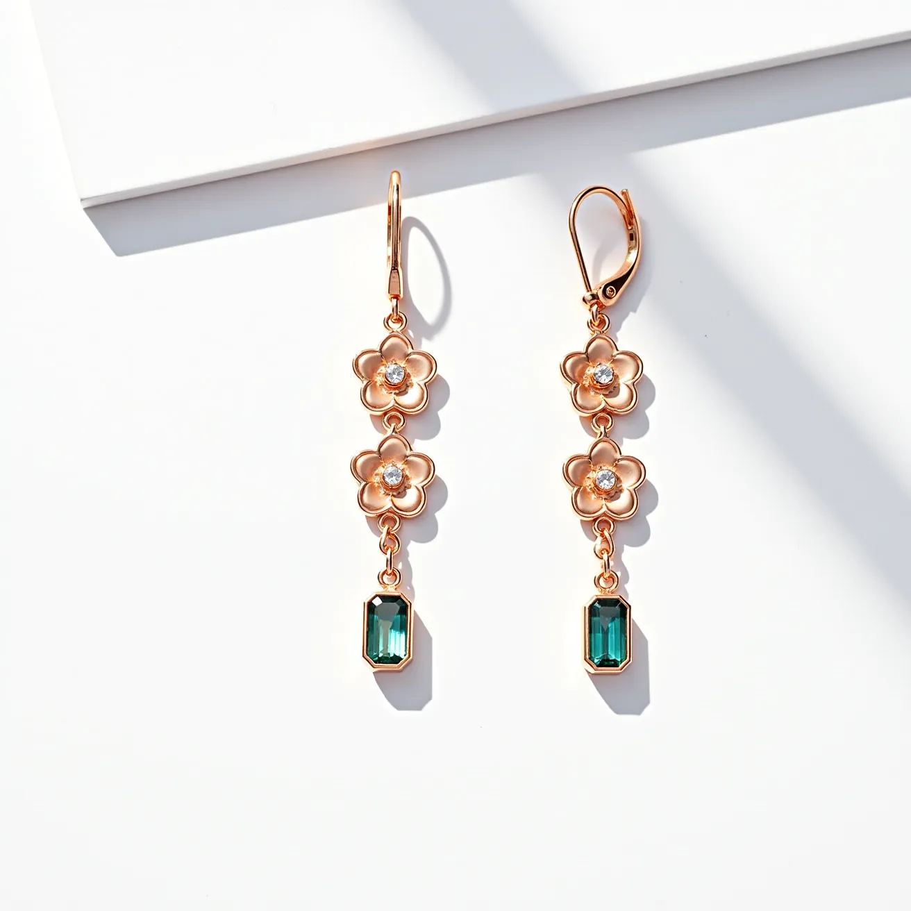 These long earrings feature a rose gold-toned metal, crafted into a delicate chain of flower motifs. Each flower exhibits a gleaming clear stone, making for an elegant setting that captures light effectively. Below these floral designs, a striking rectangular green gemstone dangles, accentuating the length of the earrings. These gemstones are bezel set, providing a clean, modern look. The earrings are secured with lever-back clasps, ensuring both style and functionality.