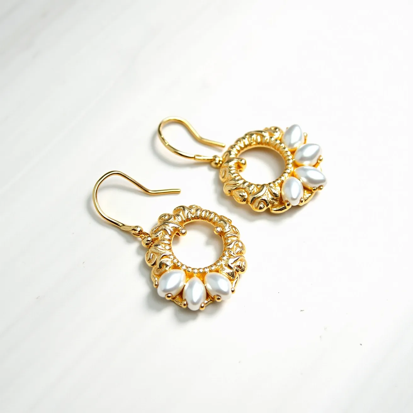 These loop earrings are crafted from a gold-toned metal, featuring intricate ornamental detailing around a circular frame. The bottom portion of each earring is adorned with three white, teardrop-shaped beads or stones, arranged in an elegant cluster. The stones are securely affixed to the frame using a prong setting, enhancing their prominence. The earrings are designed with a classic fish hook clasp, ensuring a comfortable and secure fit for the wearer.