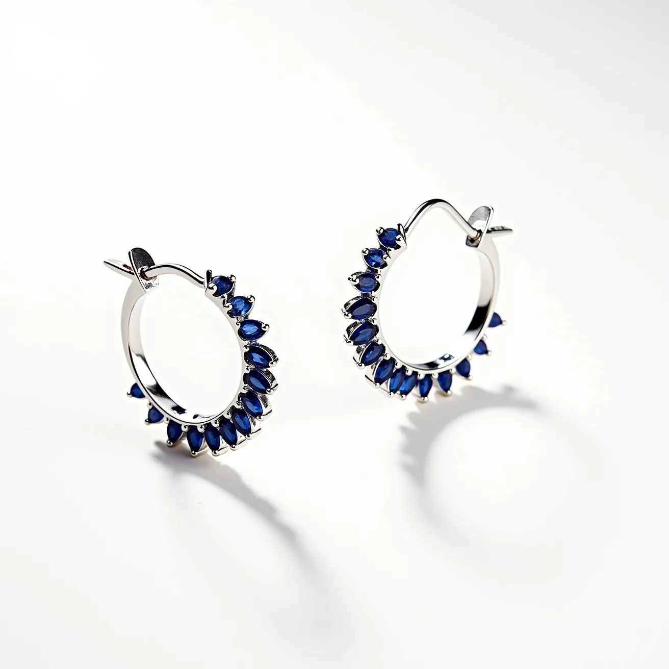 These loop earrings feature a polished silver metal that forms the primary structure of each piece, providing a sleek and elegant finish. Adorning the circumference are deep blue marquise-cut stones, likely sapphires, which are delicately prong-set to enhance their vibrant color and refractive qualities. The design of the earrings includes a classic hinged clasp, ensuring secure attachment when worn. Each stone is carefully aligned to create a captivating symmetry, making the earrings both stylish and sophisticated in appearance.