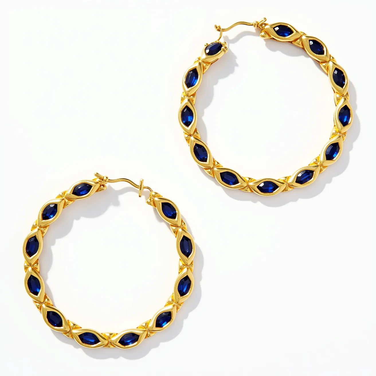 These loop earrings feature a gold-tone metal that forms the structure, providing an opulent and classic backdrop for the embedded gems. The earrings boast marquise-cut blue stones, which are consistently placed in an alternating pattern along the loop, each set within an intricate bezel-like setting that securely holds each gem in place while allowing light to enhance their rich hue. The earrings are designed with a latch-back clasp, ensuring they remain fastened comfortably and securely when worn. The combination of the gold-tone metal with the deep blue stones creates a bold and elegant aesthetic, suitable for both formal and casual occasions.