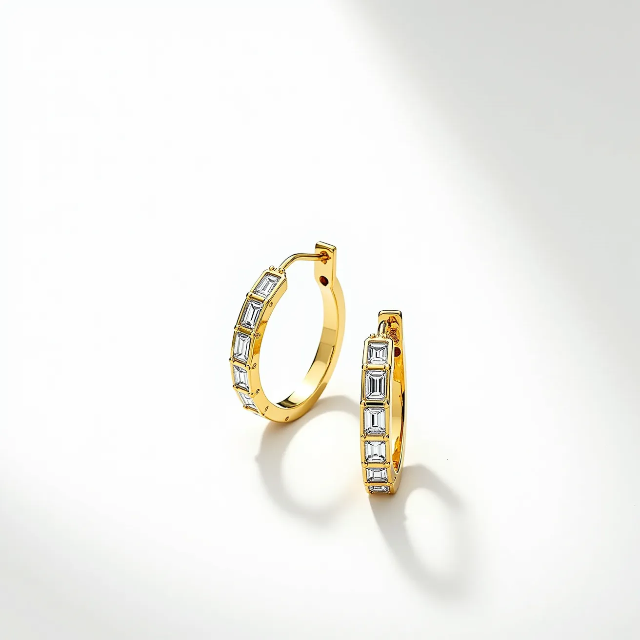 These loop earrings are crafted from a shiny gold metal, forming a smooth, circular hoop. Each earring is adorned with a series of rectangular, emerald-cut clear stones, beautifully set alongside the outer perimeter in a channel setting. The inclusion of these stones adds a touch of elegance and sparkle. The earrings feature a hinge mechanism with a secure latch back clasp, ensuring they can be worn comfortably and safely. The overall design offers a sleek and sophisticated look, making them suitable for various occasions.