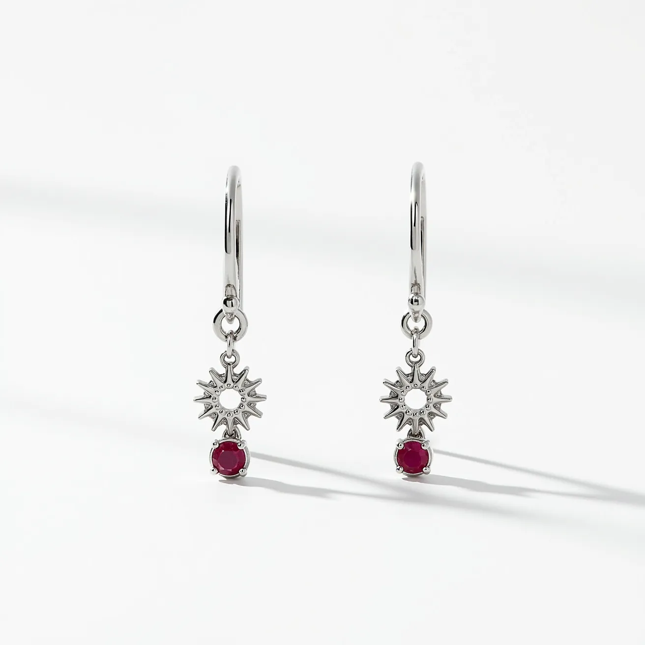 These loop earrings feature a sleek metallic hoop made from a shiny, polished material, likely silver or a silver-toned metal. Dangling from each hoop is a sun-shaped charm with intricate detailing, creating a radiant and eye-catching design. Beneath the sun motifs, there is a round, red gemstone that adds a pop of color, set in a classic four-prong setting, which securely holds the stone while allowing maximum light to enhance its brilliance. The earrings are equipped with a simple latch or snap closure, ensuring that they remain securely attached when worn.