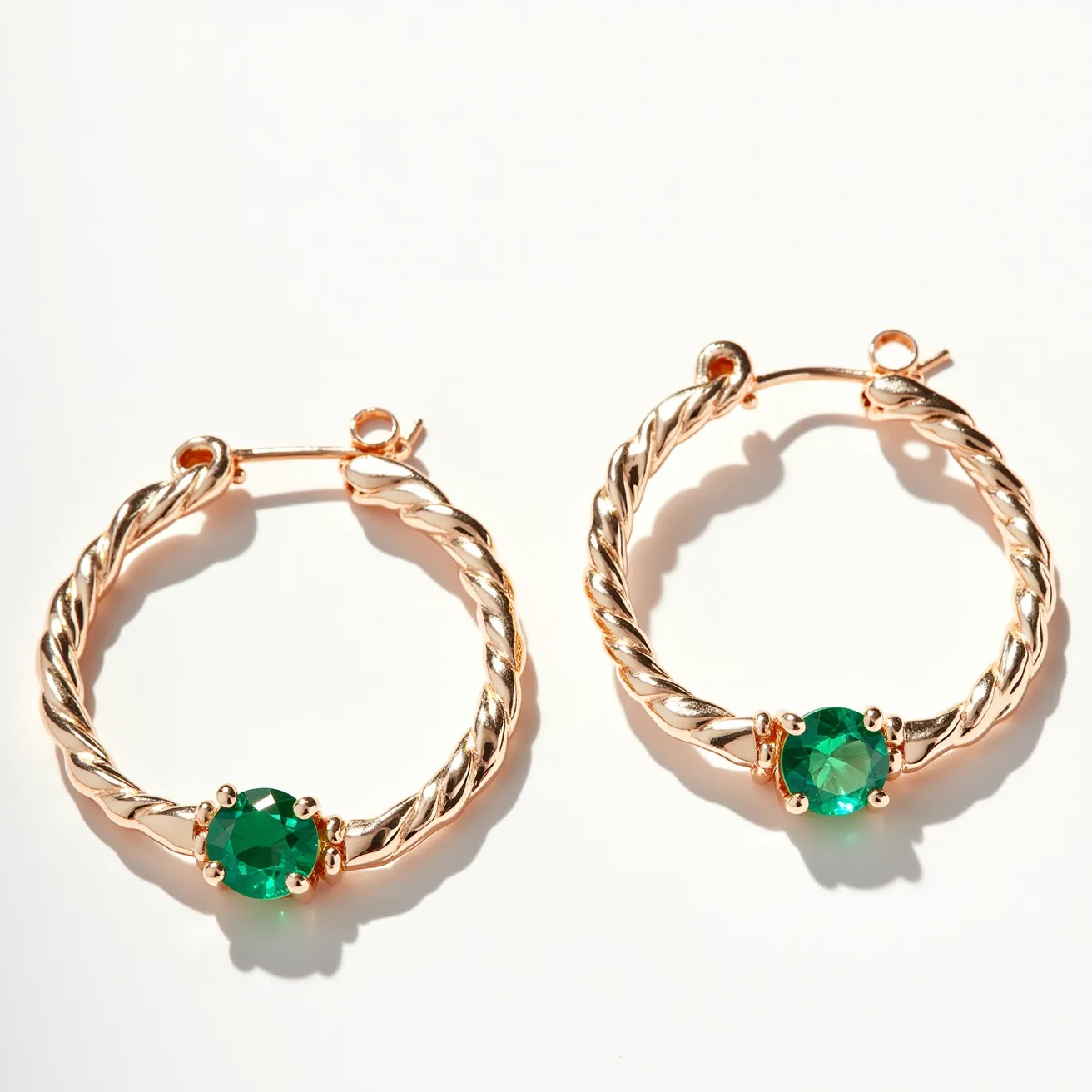 These loop earrings feature a twisted metal design, likely crafted from a gold-toned material that provides an elegant sheen. Prominently displayed at the center of each earring is a round-cut green gem, securely set in a four-prong setting. The earrings utilize a latch-back clasp for secure wear. The combination of the twisted design and the vibrant gemstone creates a striking statement piece.