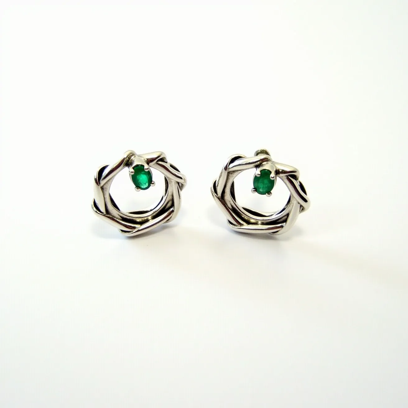 These loop earrings are crafted from a silver-toned metal featuring an artistic and interwoven design that forms a circular shape. At the center of each earring is a striking green oval gem, possibly an emerald, secured with a prong setting. The vibrant green stones add a pop of color and elegance to the overall design, while the intertwined metal loops provide a modern and sophisticated aesthetic.