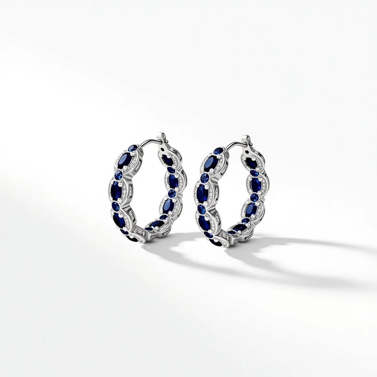 These loop earrings showcase a sophisticated design crafted from a shiny metallic material, likely silver or white gold. They are adorned with alternating oval and round gemstones, which appear to be sapphires due to their deep blue hue. Each sapphire is meticulously set in a secure prong setting, enhancing both the durability and the brilliance of the earrings. The hoop design is elegant, with a smooth curvature that adds a timeless and classic touch. A hinged snap back clasp provides a secure and comfortable fit, ensuring the earrings are both stylish and functional.