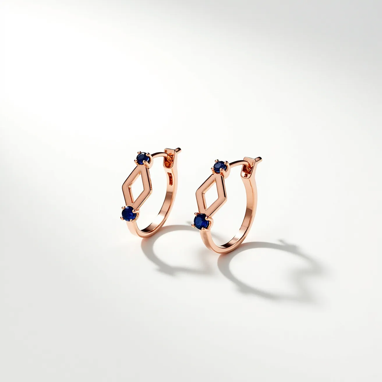 These loop earrings feature a delicate hoop crafted from a lustrous rose gold material. Each earring is adorned with two striking deep blue round-cut gems, likely sapphires, that are securely set in a classic prong setting. The design incorporates a geometric diamond shape as part of the hoop, adding an elegant and modern touch. The earrings are fastened with a hinged clasp, ensuring a secure and comfortable fit for the wearer.