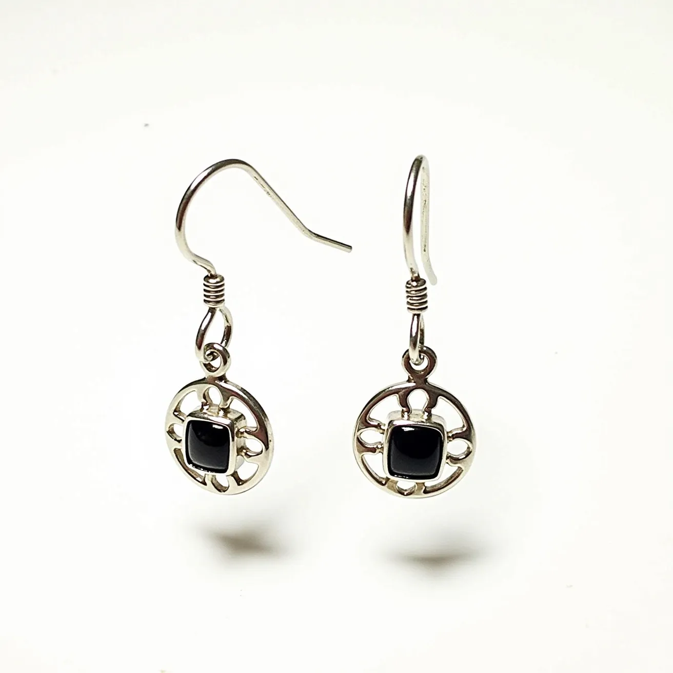 These men's dangle earrings feature an elegant design composed of a circular metal framework, likely silver, which serves as the base for each earring. At the center of the circular design sits a small, square-cut black stone, likely onyx or a similar dark gem, securely held in a bezel setting. The metal framework surrounding the stone includes intricate cut-out patterns, adding to the visual appeal. The earrings are attached with traditional fish hook clasps, providing a simple yet secure attachment for wearing. The combination of the dark stone and silver-toned metal creates a striking contrast, making these earrings a distinctive accessory.