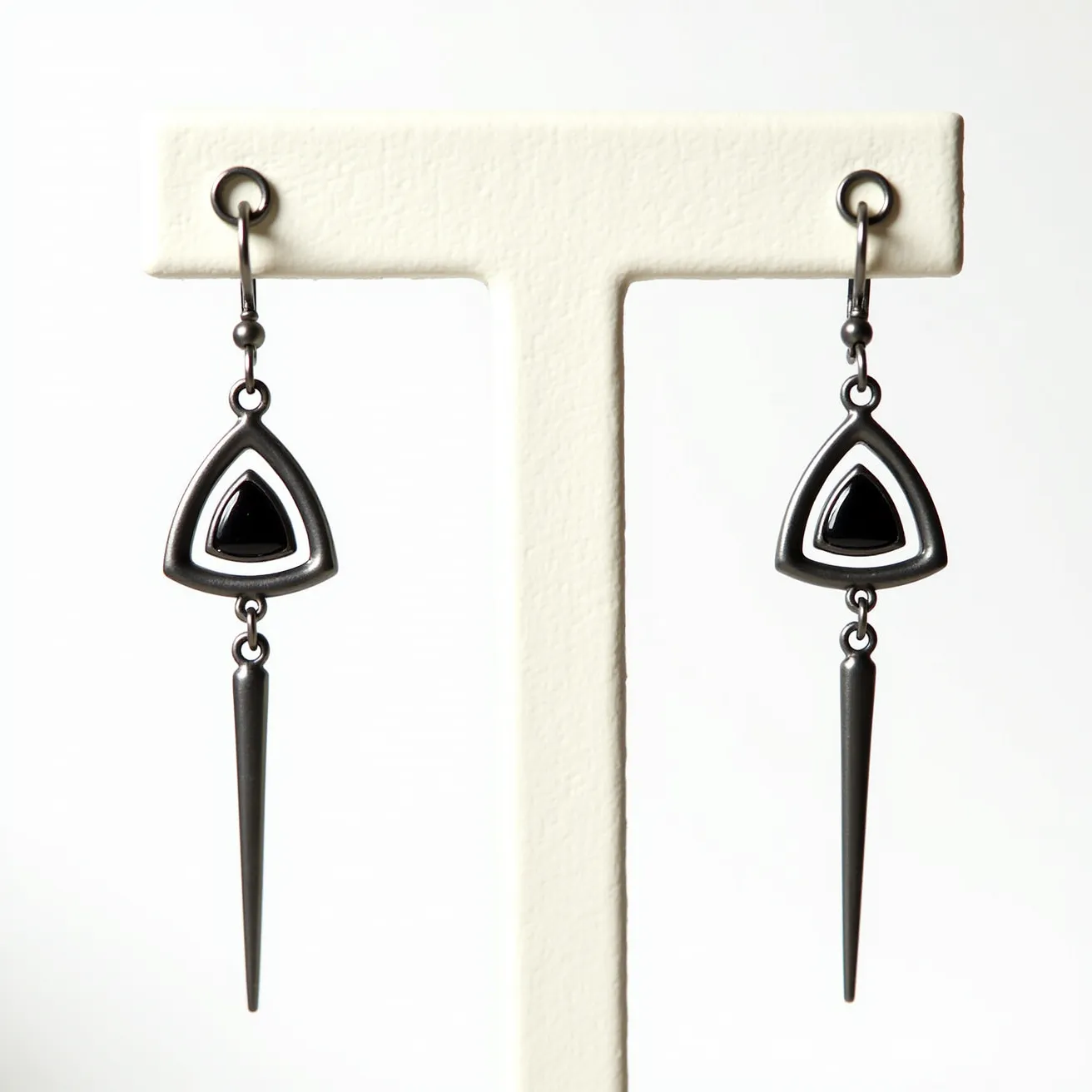 These men's dangle earrings feature a modern design with triangular elements and pointed drops. The earrings appear to be crafted from a dark metal, likely stainless steel, giving them a sleek, contemporary look. Each earring showcases a central triangular black stone or enamel inset, which is bordered by a matching triangular metal frame, creating an eye-catching geometric effect. The stone is likely cabochon cut, providing a smooth surface and glossy finish. The sharp, elongated cone dangles beneath, adding a touch of edginess to the design. The earrings attach with a hoop and small latch clasp, ensuring a secure fit while maintaining their stylish appearance.