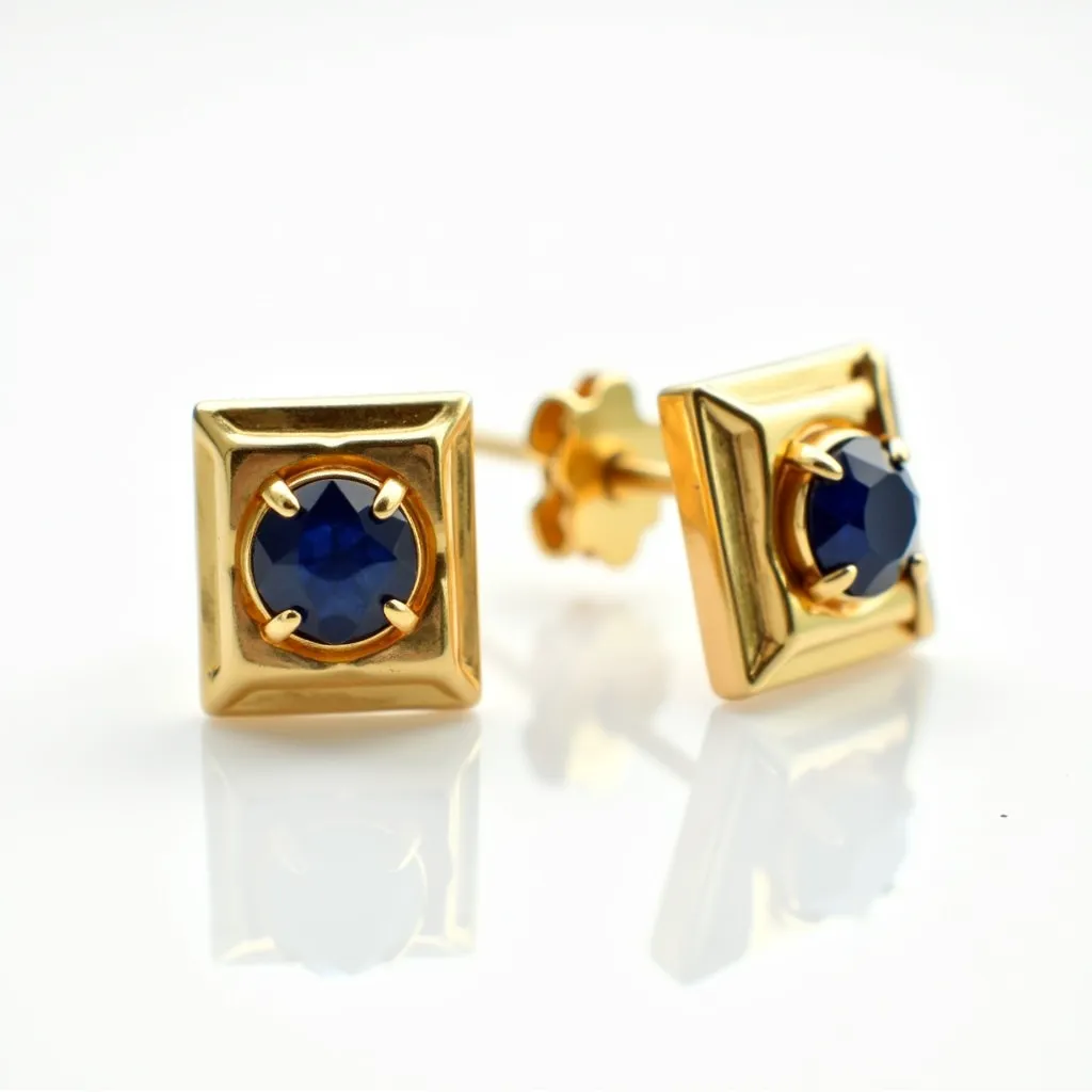 These men's earrings feature a bold design with a square-shaped gold setting. The center showcases a round, deep blue gemstone, possibly a sapphire, held securely by four prongs, emphasizing its elegant cut. The gemstone is nestled in a recessed frame, enhancing its prominence against the gold backdrop. The earrings are equipped with a push-back clasp mechanism, ensuring secure and comfortable wear. The overall aesthetic balances a classical approach with a touch of sophistication, making them a versatile accessory.