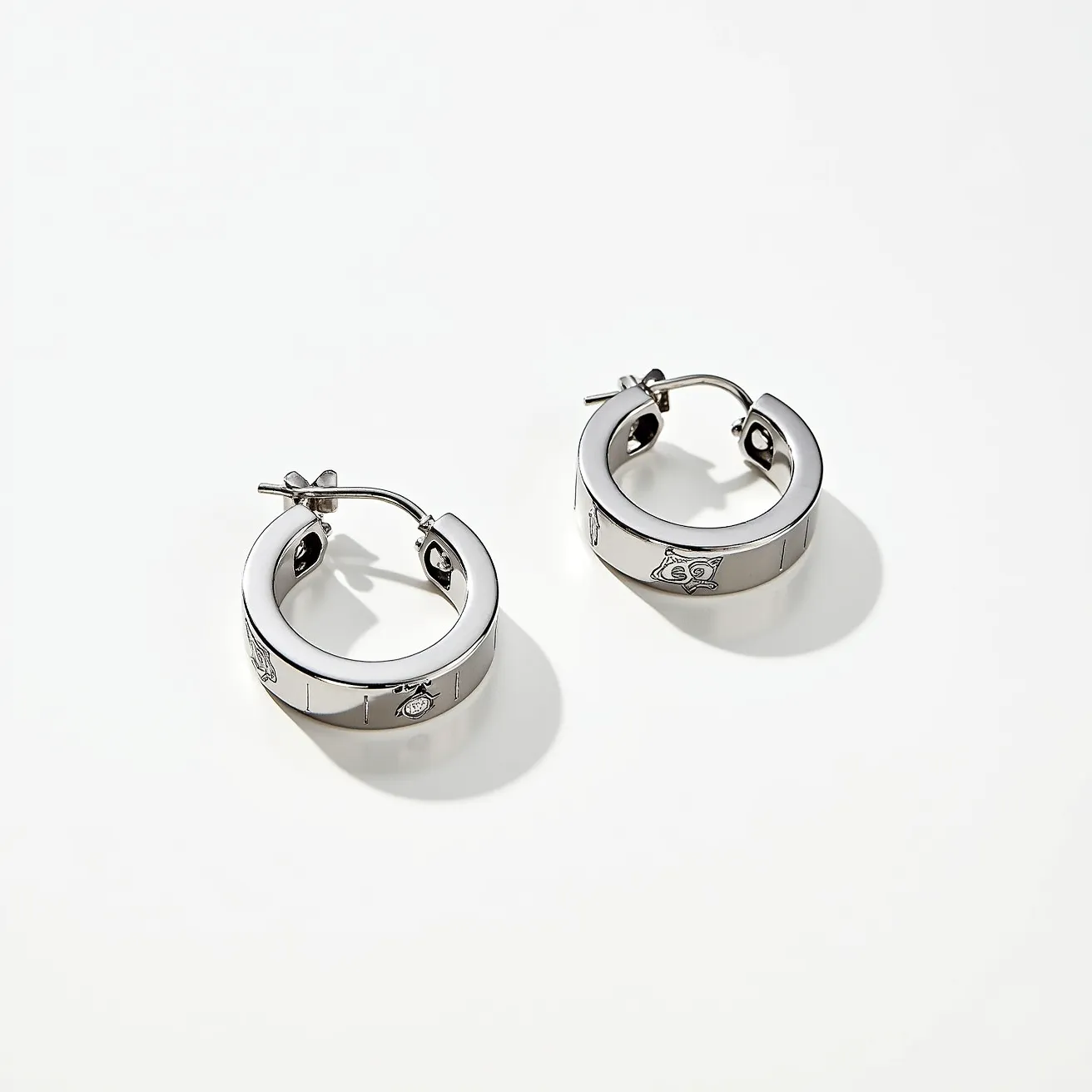 These men's hoop earrings are crafted from a polished metal, likely stainless steel, giving them a sleek and modern appearance. The surface features engraved emblems that add decorative interest to the design. The hoops are relatively thick, contributing to a bold and substantial look. They are equipped with a hinge and latch clasp, ensuring secure and comfortable wear. The design is unembellished by additional stones or gems, keeping the focus on the lustrous finish and subtle engravings.