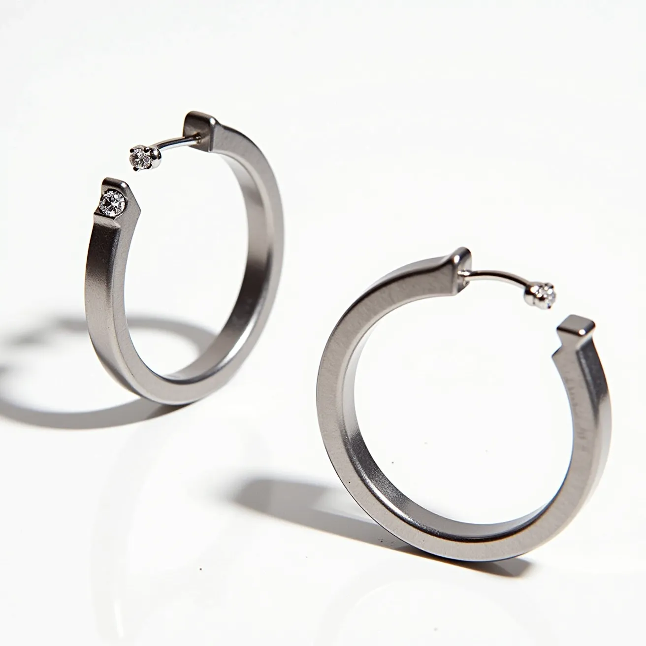 These men's hoop earrings feature a sleek, modern design crafted from a polished metal, likely stainless steel, giving them a robust and contemporary appearance. Each earring is adorned with a small, round-cut stone, which is set at one end of the hoop, adding a subtle yet elegant touch. These stones appear to be securely and neatly set, enhancing their sparkle and presence. The earrings are designed with a hinge clasp mechanism, ensuring a secure and comfortable fit when worn. The overall design is both stylish and understated, suitable for both casual and formal occasions.