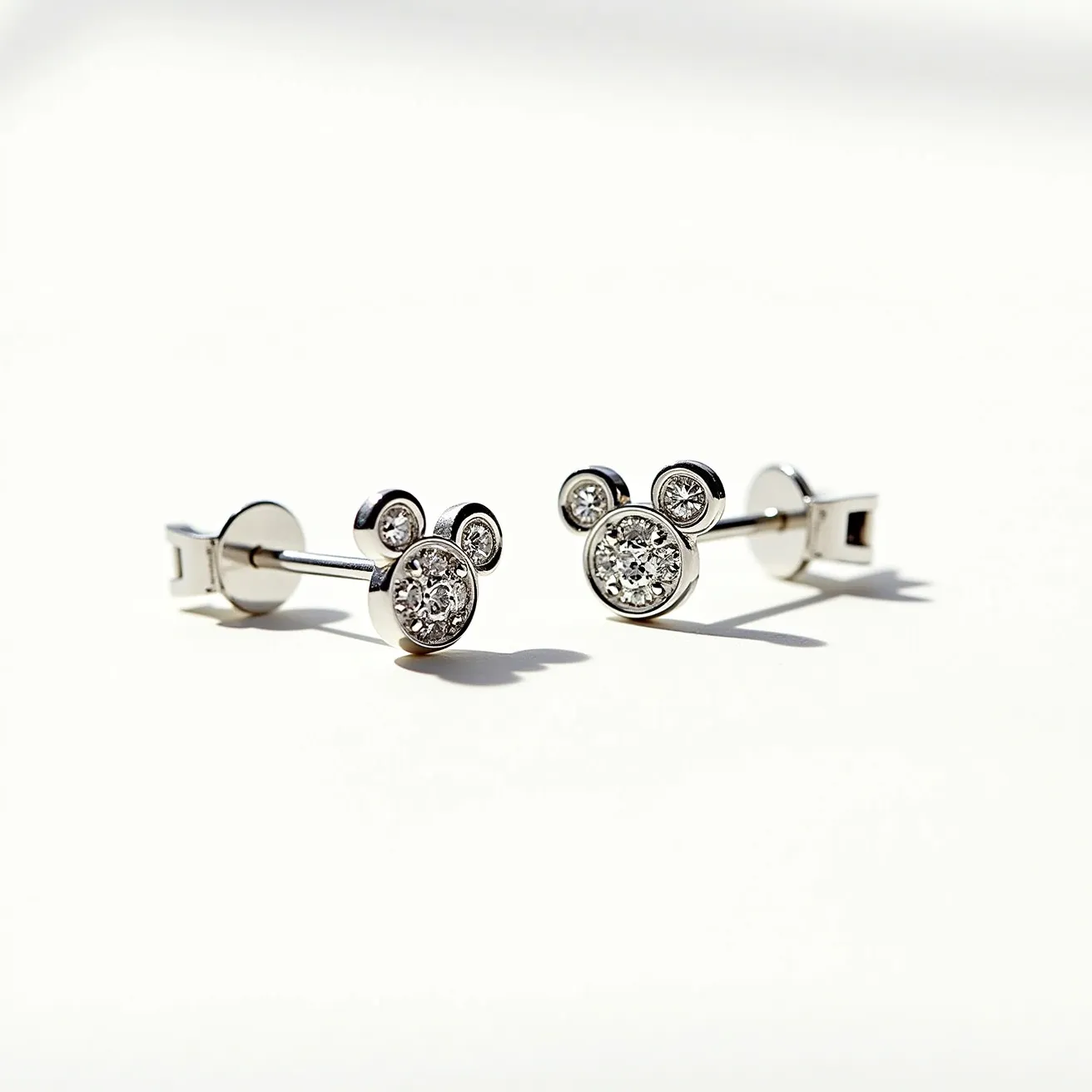 These mickey mouse earrings feature a classic design with a bold silhouette that represents the beloved cartoon character. Crafted from polished metal, likely silver or white gold, these earrings present a sleek finish. The central component of each earring showcases three clear round-cut stones—potentially diamonds—set in a flush style that forms the iconic mouse head shape. The stones are uniformly set, ensuring a balanced and symmetrical appearance. These earrings utilize a secure post and butterfly clutch for attachment, providing both style and reassurance to the wearer.