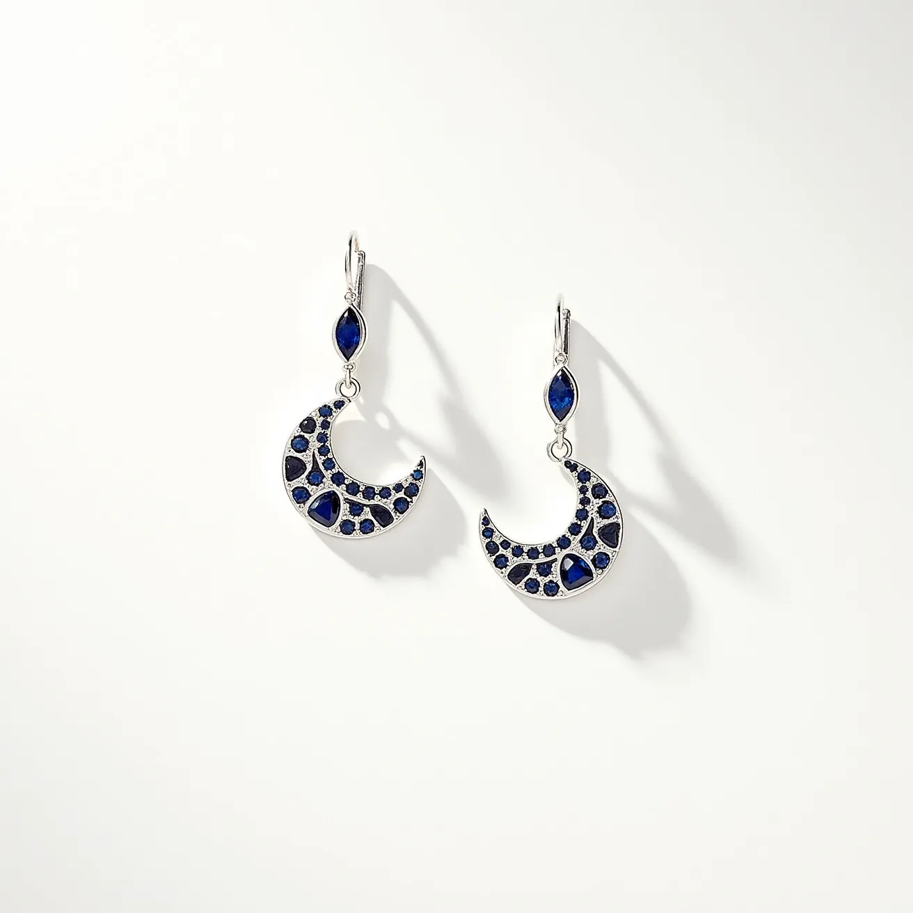 These moon earrings feature a crescent shape adorned with deep blue gemstones, lending them an elegant and celestial appearance. The gems appear to be round and teardrop cuts, set in a structured pattern that highlights the moon motif. The metal seems to be a polished silver or a similar alloy, providing a sleek and reflective backdrop to the stones. The earrings are connected to the ear with a secure lever-back clasp, ensuring they stay firmly in place while adding a touch of sophistication to their design. The combination of the cooler silver tone with the rich blue gemstones creates a striking yet harmonious effect, evoking the serene beauty of a night sky.