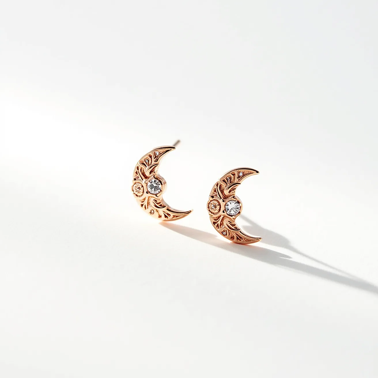 These moon earrings feature a crescent shape crafted from a warm, golden metal, possibly rose gold, which gives them a delicate and luxurious appearance. Intricate detailing on the surface of the crescents adds an elegant, ornate look. Each earring is adorned with a single, central gemstone, likely a clear crystal or diamond, set in a bezel setting for a secure and refined finish. The posts indicate a simple push-back fastening system, providing ease of wear while maintaining their elegant design. The overall aesthetic combines celestial motifs with classic elegance, making these earrings a versatile accessory.