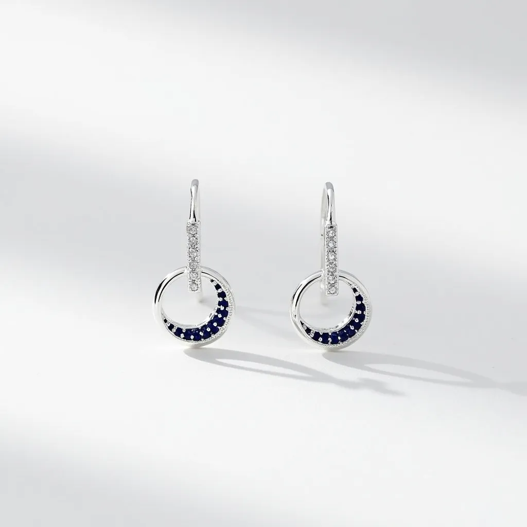 These moon earrings feature silver-toned circular pendants with a delicate integration of blue and white stones. The blue stones, possibly sapphires, are set in a pavé style, creating a crescent shape within the circular design, elegantly outlined by smaller clear stones that add sparkle and contrast. The earrings are attached to a hook clasp that extends into a long, slender drop, adorned with additional white stones, providing a seamless and cohesive connection between the earring elements. The arrangement and choice of materials give these earrings a celestial elegance, perfectly emulating the mystique and allure of the moon.