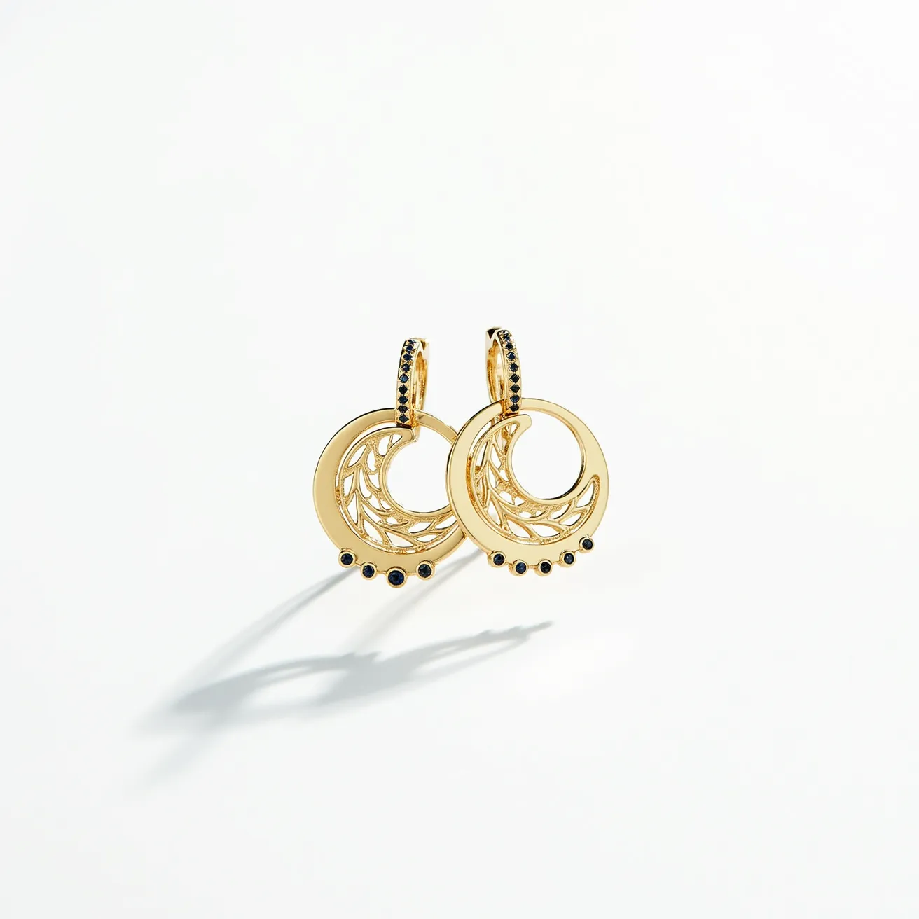 These moon earrings feature a circular design crafted from a gold-toned metal, showcasing a delicate, crescent moon pattern within the circle. The outer rim of each earring is adorned with small black gemstones, likely set in a bezel setting, offering a subtle contrast against the gold. The earrings are attached via a small hoop mechanism, which also appears to be embellished with matching black stones, ensuring both aesthetic and secure wear. The overall design combines elegance with a modern twist, achieved through the intricate detailing and shimmering accents.