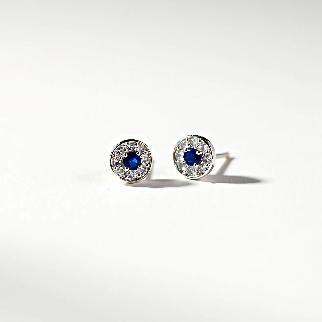 These moon earrings feature a central blue gemstone, likely a sapphire, which is round-cut and set in a bezel style. Surrounding the central stone is a circle of smaller, clear stones that resemble diamonds, set in a pavé setting to enhance their sparkle. The earrings are crafted from a silver-toned metal, potentially white gold or sterling silver, providing a sleek and polished appearance. They are equipped with post attachments, suitable for pierced ears, ensuring secure fit and comfort. The design balances the rich, deep hue of the blue stone with the brilliance of the clear stones for an exquisite celestial look.