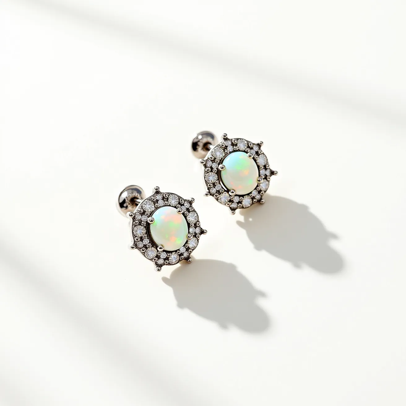 These opal earrings feature an elegant design with central opals exhibiting a captivating play of color. The opals are oval-cut and are set in a halo style, surrounded by shimmering, small round-cut diamonds or diamond-like stones, enhancing their brilliance. The setting appears to be crafted from a metal such as white gold or platinum, adding a refined touch to the overall aesthetic. The earrings are finished with a secure post and butterfly clasp, ensuring they remain comfortably and safely in place when worn.