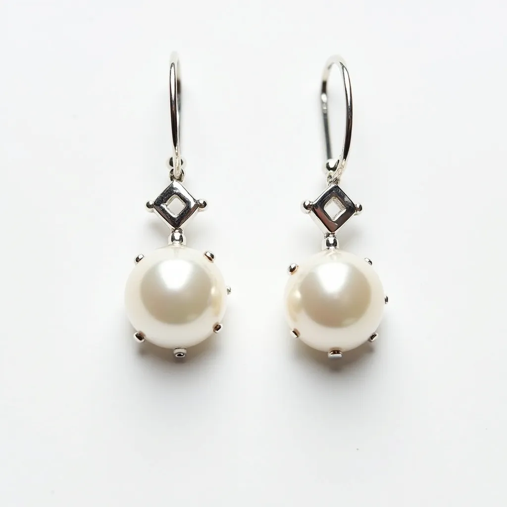 These pearl drop earrings feature elegant round pearls, set in a lustrous silver-toned metal that complements their natural shine. Each pearl is held securely by a series of prongs, which add a subtle decorative element to the design. Above the pearls, there is a geometric square detail, also crafted in the same metal, adding a modern touch to the classic look. The earrings are designed with a simple hook clasp, allowing for easy wear while maintaining a graceful aesthetic.