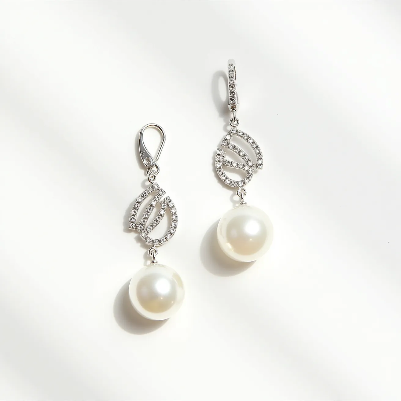 These pearl drop earrings feature an elegant design with round, lustrous pearls hanging from an intricate metal setting. The metal, likely silver or white gold, is adorned with small, shimmering gemstones, possibly diamonds, arranged in delicate loops. The gemstones are round cut and pave set, providing a subtle sparkle that complements the pearls. Each earring is secured with a lever-back clasp, ensuring a comfortable and secure fit. The overall design is both sophisticated and timeless, making these earrings suitable for both formal occasions and elegant everyday wear.