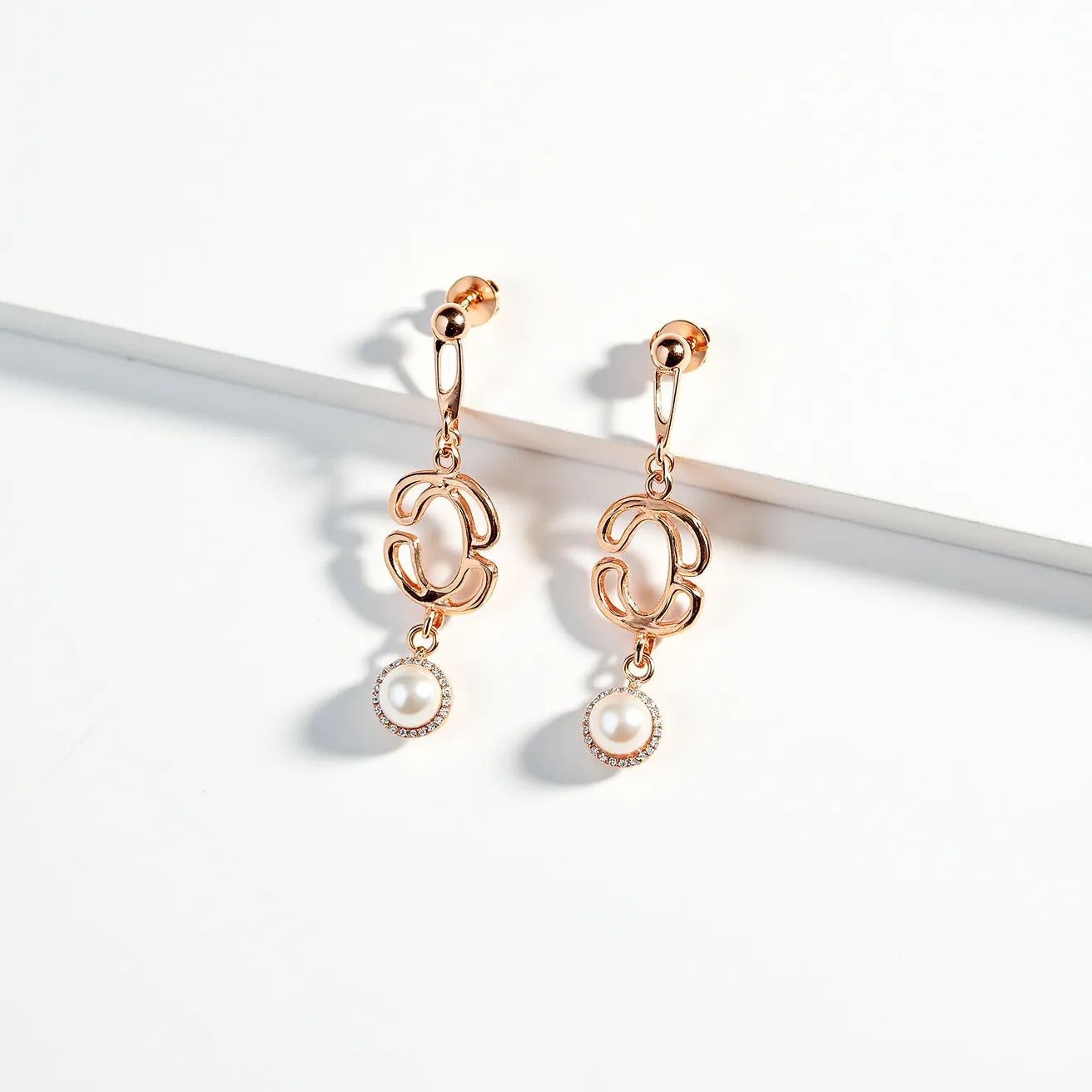 These pearl drop earrings feature a refined combination of sleek gold metal and lustrous pearls. The design showcases a unique motif, likely crafted from gold-plated metal, which serves as a central element in the structure. Suspended from the motif is a single pearl, each encircled by a delicate halo of small, sparkling gemstones that accentuate the pearl’s elegance. The earrings are attached with a post and butterfly clasp, ensuring a secure and comfortable fit. This elegant piece exemplifies classic sophistication with a modern twist, making it suitable for both formal and casual occasions.