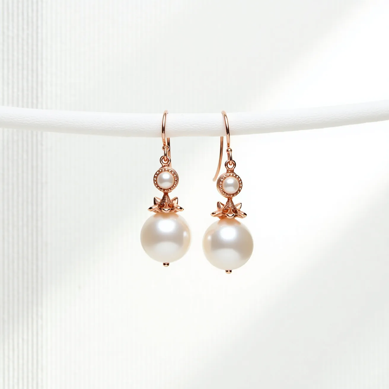 These pearl drop earrings feature lustrous, round pearls as the main focal point, elegantly suspended beneath smaller, bead-set pearls encased in a decorative gold setting. The intricate design includes a floral-like metal accent above the larger pearls, providing an additional touch of sophistication. The earrings are crafted with a gold hook clasp that ensures secure and comfortable wear, highlighting a harmonious blend of classic elegance and modern design. The materials and craftsmanship reflect an attention to detail that enhances the natural beauty of the pearls.