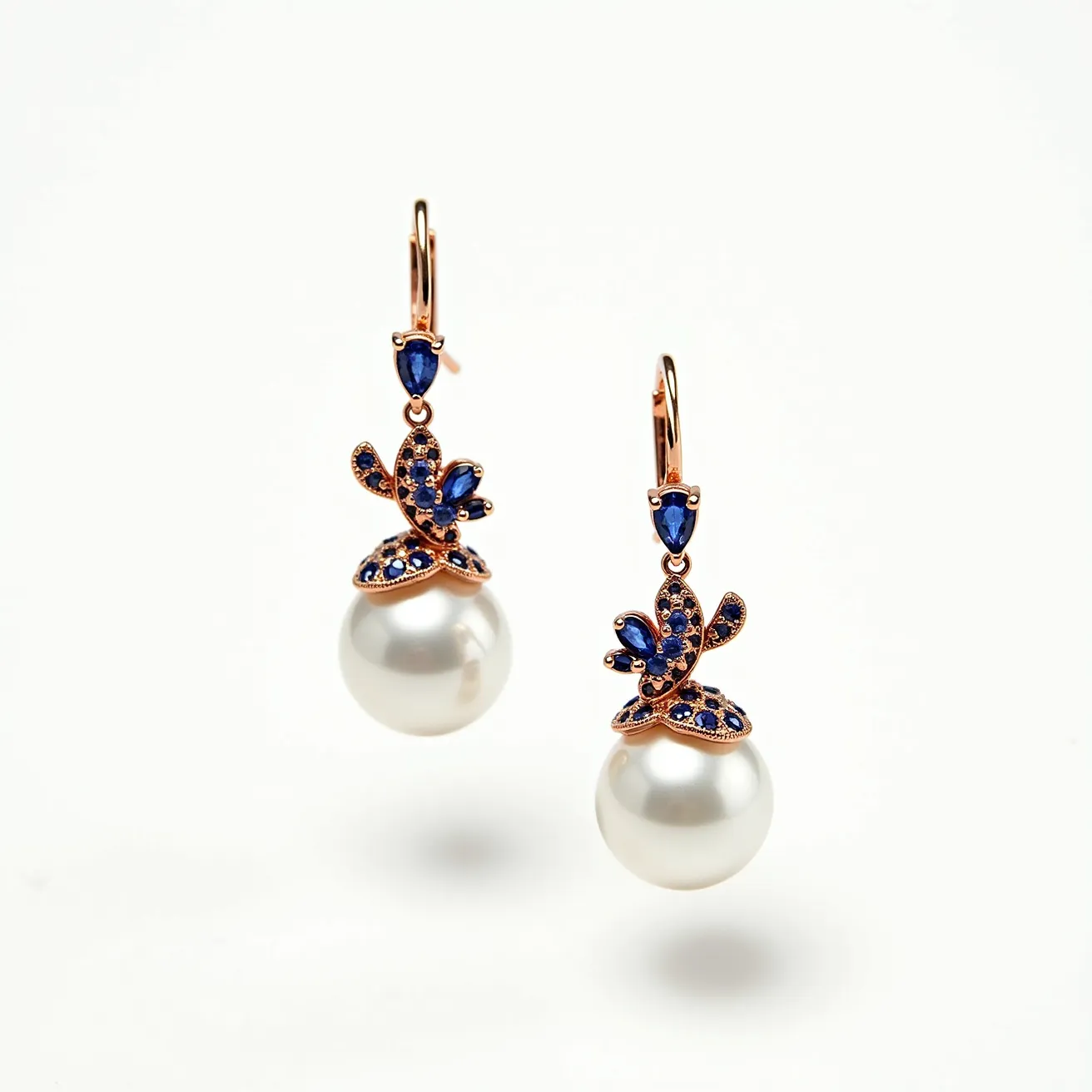 These pearl drop earrings feature a stunning combination of lustrous white pearls and vibrant blue gemstones, likely sapphires, set in a delicate, intricate pattern. Each earring showcases a large, round pearl that gracefully hangs from a gold or gold-toned setting, adding a classic elegance to the piece. Above the pearls, the blue gemstones are artfully arranged in a floral motif, with pear and round cuts that catch the light beautifully. The gemstones are secured in prong settings, adding both stability and an aesthetic appeal. The earrings are equipped with a lever back clasp, ensuring a secure and comfortable fit when worn.