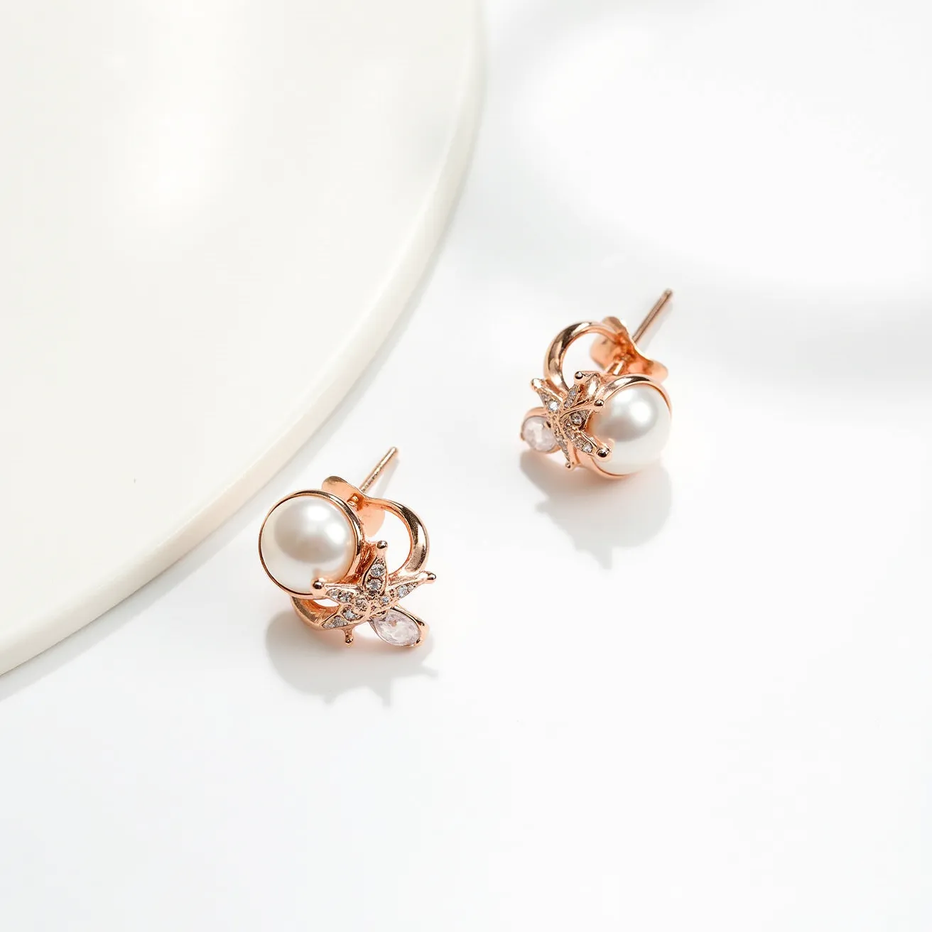 These pearl earrings feature a central, lustrous round pearl set in a rose gold setting. Surrounding the pearl is a delicate starfish motif adorned with small, clear gemstones that are possibly diamonds or cubic zirconia, adding a sparkling accent. A small, teardrop-shaped gemstone is positioned strategically beneath the starfish design, enhancing the elegance of the piece. The earring is attached with a simple post-back clasp, providing ease of wear while maintaining secure placement on the ear. The combination of pearls, rose gold, and gem embellishments creates a sophisticated and stylish accessory.