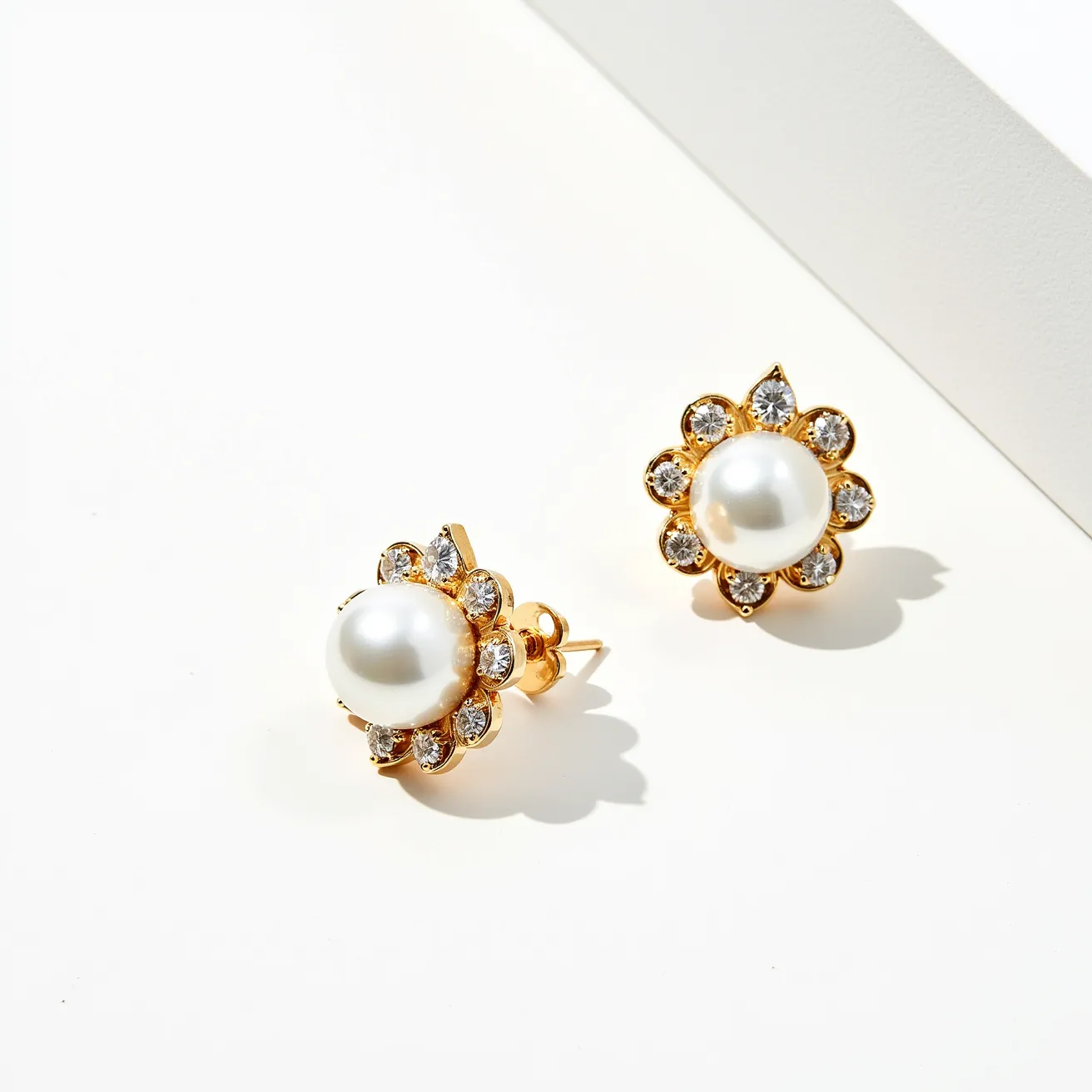 These pearl earrings feature a classic and elegant design, centered around a smooth, lustrous white pearl. Surrounding the pearl is a floral-like arrangement of clear gemstones, likely diamonds, each set in a prong setting, enhancing the earrings’ brilliance and adding a touch of sparkle. The earrings are crafted from a gold-toned metal, which provides a warm and rich contrast to the cool sheen of the pearls and gems. They are designed as stud earrings, with a secure post and butterfly clasp, ensuring they stay comfortably in place when worn.