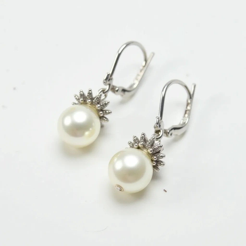 These pair of pearl earrings features elegant round pearls as the central gem, displaying a classic and lustrous appearance. The pearls are complemented by a decorative setting that resembles a floral or sunburst motif, crafted in a metallic finish that suggests a platinum or white gold material. Small, inset stones—likely diamonds—accentuate this motif, adding a subtle sparkle to the overall design. The earrings are secured with lever-back clasps, offering both style and functionality, ensuring that the earrings remain comfortably in place when worn.