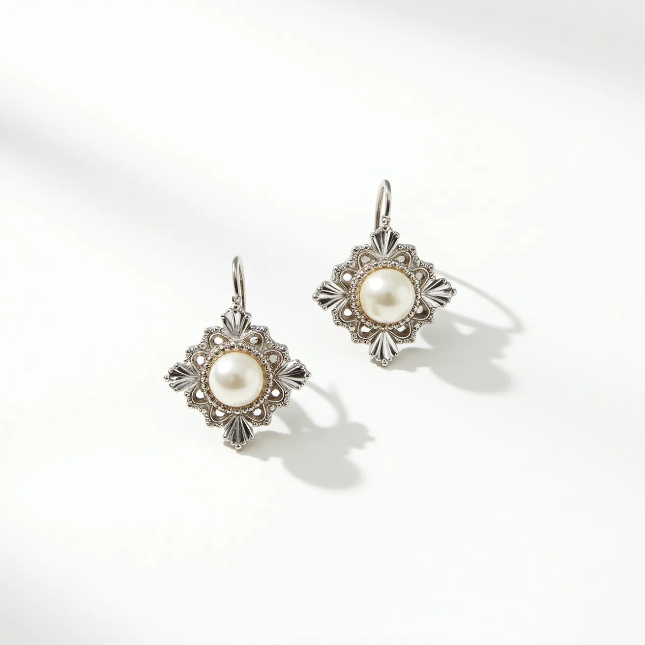 These pearl earrings feature round pearls set as the focal point, surrounded by intricate silver-toned metal work that highlights their classic elegance. The pearls are mounted in a bezel setting, enhancing their smooth luster and natural beauty. The decorative metal elements exhibit an ornate design reminiscent of vintage styles, adding a touch of timeless sophistication. These earrings utilize a hook attachment, allowing them to dangle gracefully and ensuring ease of wear. The combination of the lustrous pearls and the detailed metal work make these earrings a standout accessory.