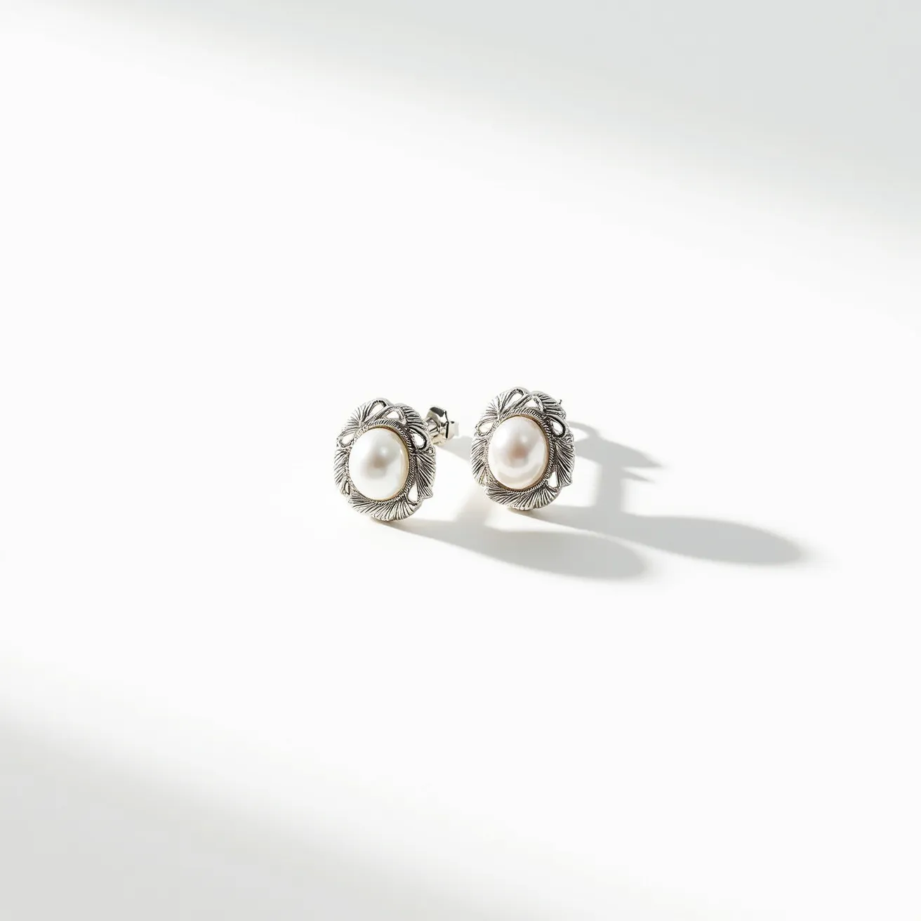 These pearl stud earrings feature lustrous, round pearls set at the center, embraced by an intricate metal setting that resembles petals or leaves, adding a touch of elegance and texture. The material surrounding the pearls appears to be silver-toned, enhancing the overall sophisticated look. The earrings utilize a classic post-back clasp for secure attachment. This design combines the timeless appeal of pearls with the detailed artistry of the metal setting, creating a versatile accessory suitable for various occasions.