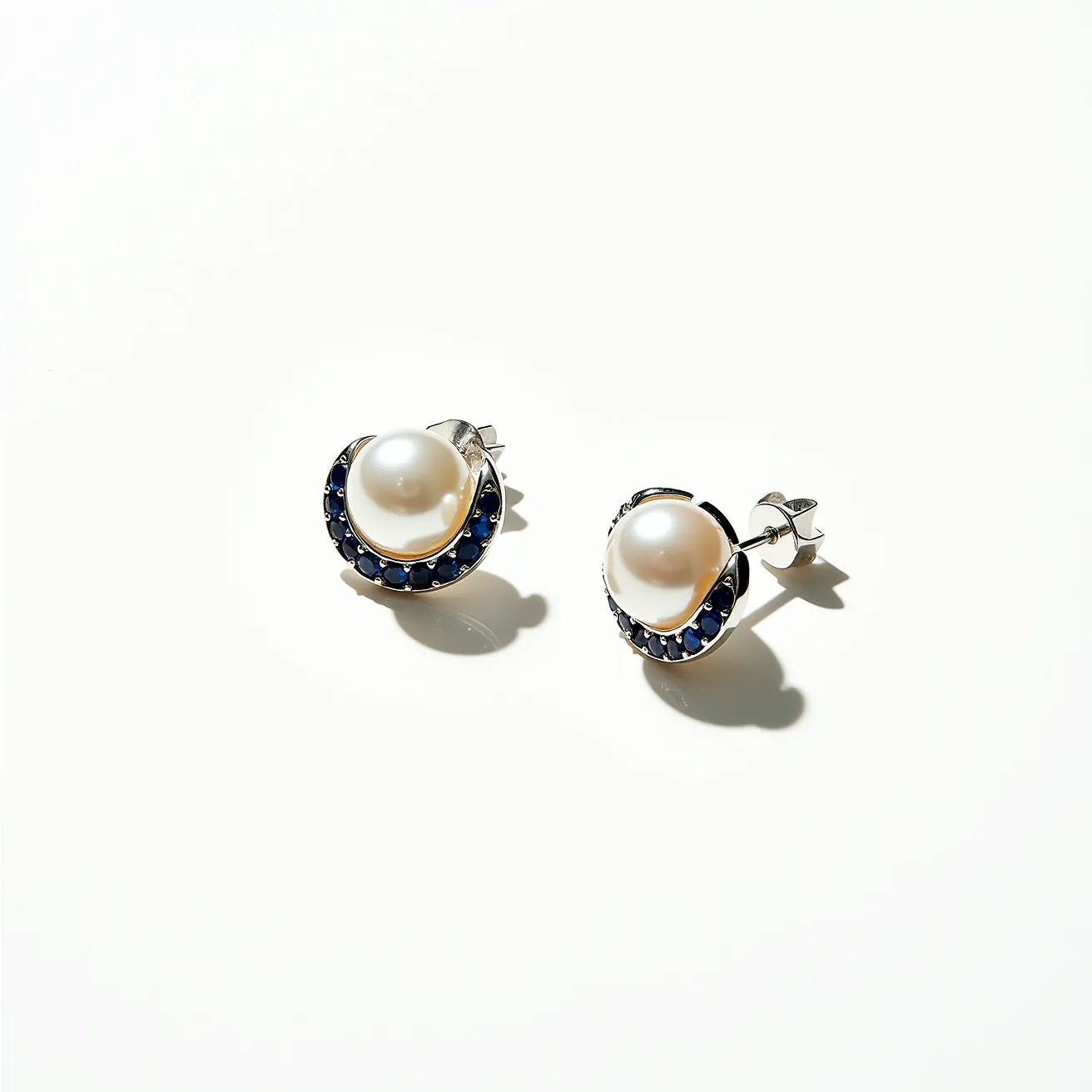 These pearl stud earrings feature lustrous pearls set at the center, surrounded by a crescent of small, round-cut blue gemstones that add a touch of color and elegance. The earrings are crafted with a silvery metal, likely silver or white gold, which complements the pearls and gemstones beautifully. They are designed with a classic post and butterfly clasp attachment, ensuring a secure fit while being easy to wear. The combination of pearls and blue stones creates a sophisticated and timeless design, suitable for various occasions.