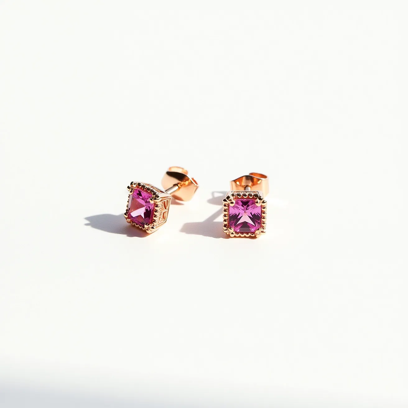 These pink earrings feature square-cut pink stones that are set in a decorative bezel setting, adding an elegant touch to their appearance. The stones are likely made from a gemstone or a high-quality synthetic material, providing a vibrant and vivid pink hue. The metal surrounding the stones appears to be a polished material, possibly gold or a gold-toned alloy, enhancing the overall luxurious feel. The earrings are equipped with a classic post-back clasp, allowing for secure and comfortable wear. The intricate detailing around the setting highlights the craftsmanship and adds a touch of sophistication.