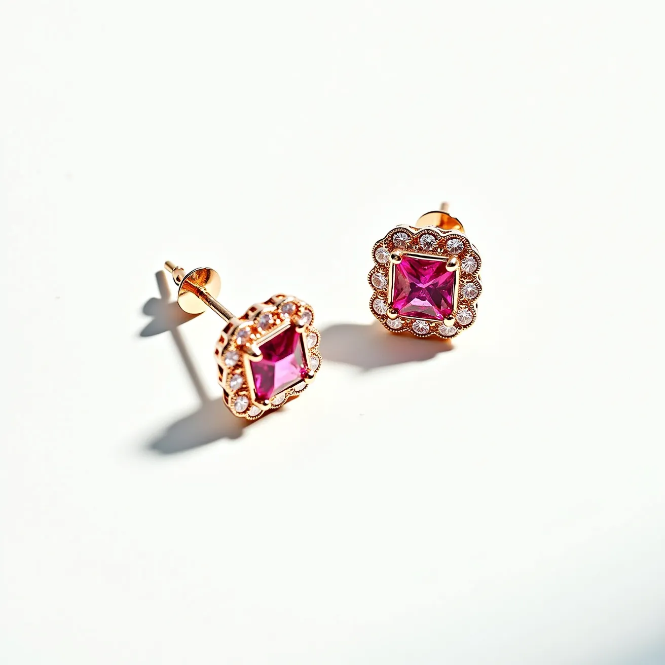 These pink sapphire earrings feature a vibrant, square-cut pink sapphire at the center, set in a secure four-prong setting. Surrounding the central gemstone is a halo of smaller white stones, likely diamonds, arranged in a scalloped design that enhances the earrings' overall elegance. The materials appear to include a polished metal, possibly white or rose gold, which complements the gemstones beautifully. These earrings use a classic stud mechanism with a push-back clasp for secure attachment to the ear, combining both style and functionality.