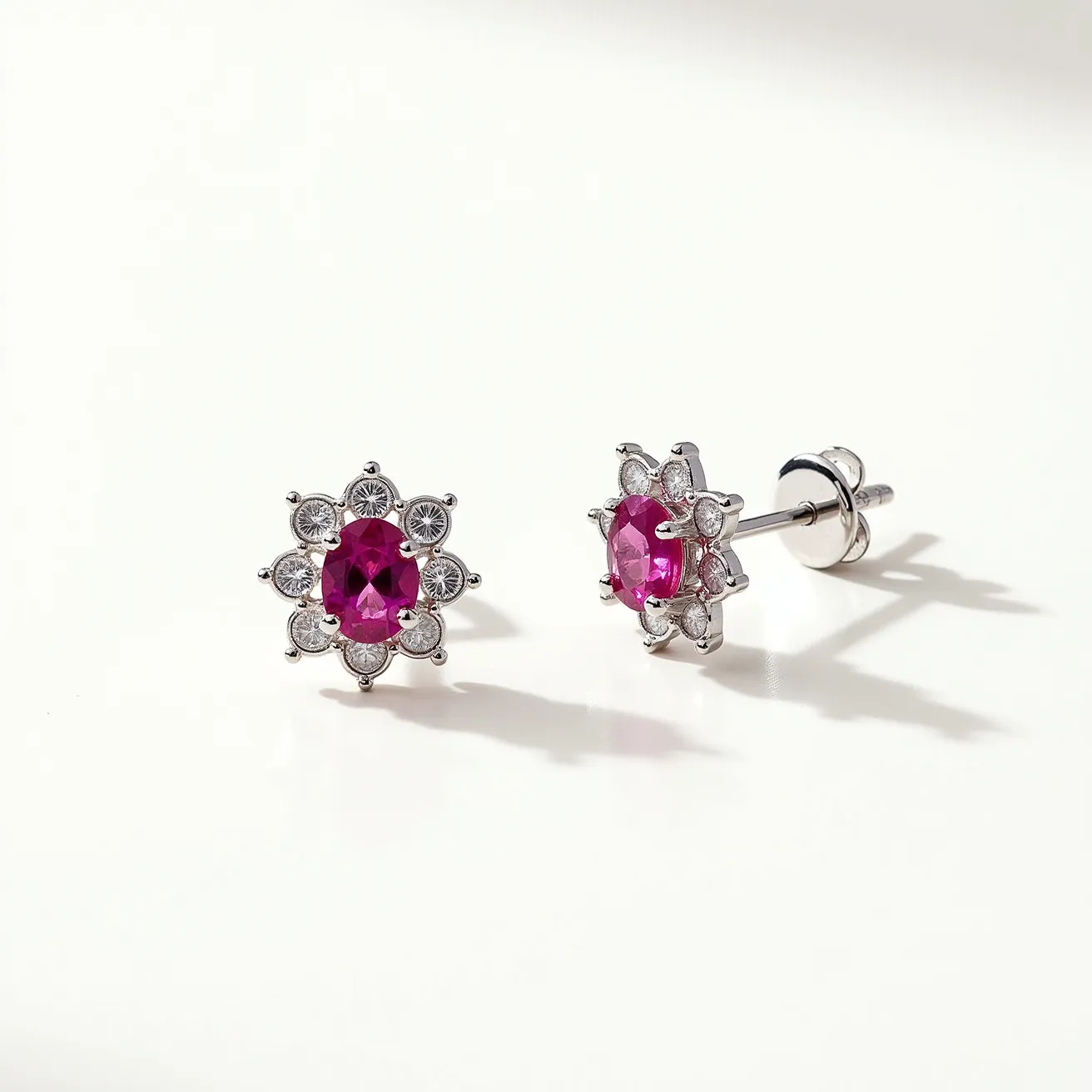 These pink sapphire earrings feature a central, oval-cut pink sapphire encircled by round white gemstones, likely diamonds, creating an elegant floral design. The setting appears to be crafted from a silvery metal, possibly white gold or platinum, which complements the gemstones beautifully. The earrings have a secure post-back closure, ensuring they stay comfortably in place. The intricate arrangement of the surrounding stones adds a touch of sophistication and enhances the vibrant hue of the central sapphires.