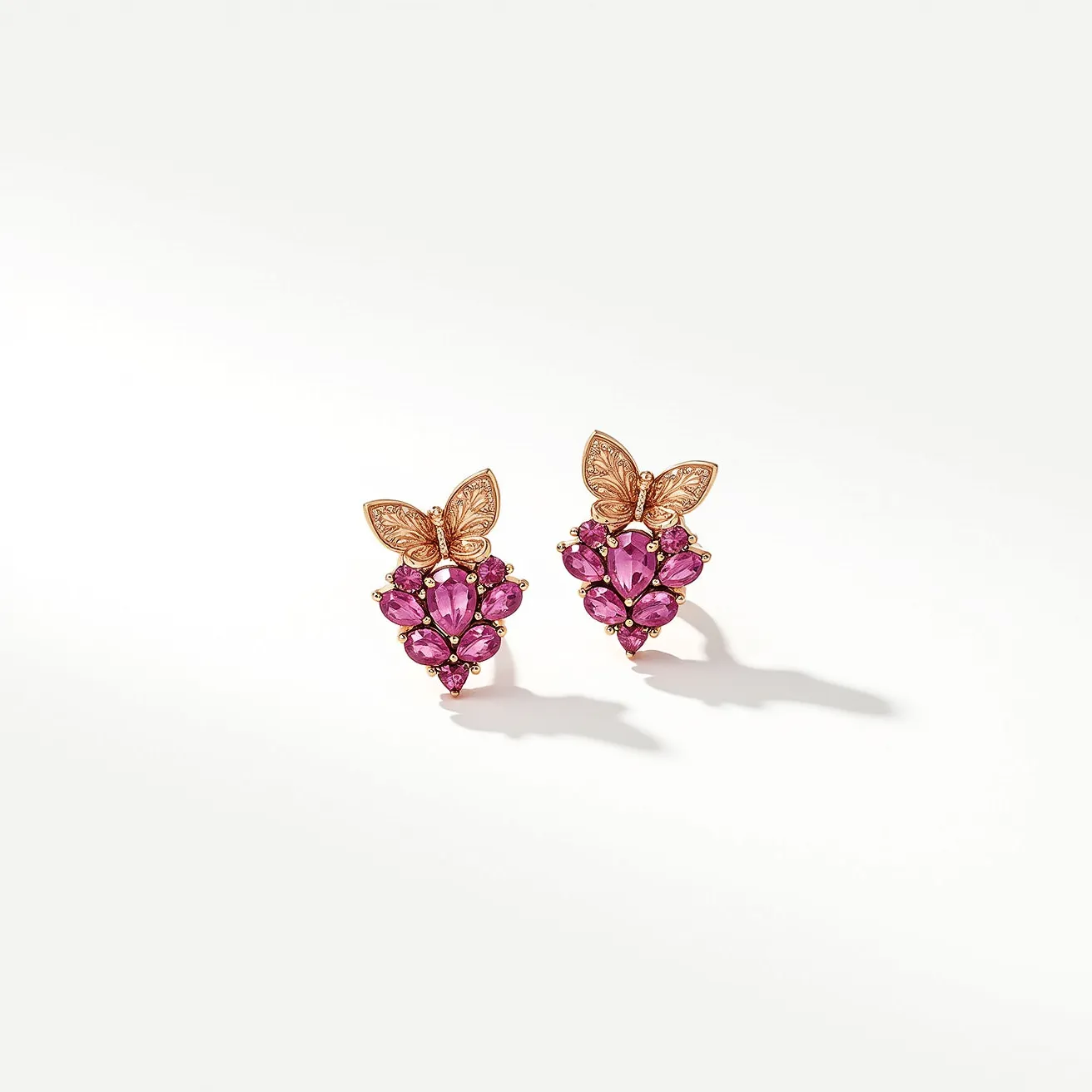 These pink sapphire earrings feature a stunning arrangement of vibrant pink sapphire gemstones set in a refined metal setting that resembles rose gold. The sapphires are skillfully cut into a combination of marquise and round shapes, forming a decorative cluster. The marquise-cut sapphires create an elegant teardrop pattern, while small round stones accentuate the design. At the top of each earring, a beautifully detailed butterfly motif adds a whimsical touch, crafted from the same metal as the setting. The earrings are fitted with secure post backings for comfortable wear.