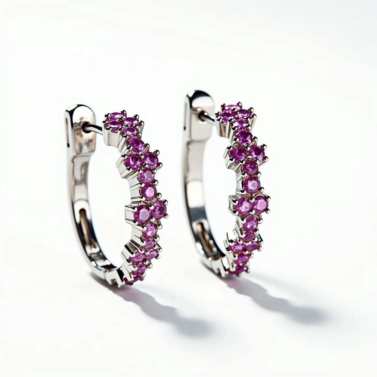 These pink sapphire earrings feature a series of vivid round-cut pink sapphires intricately set along the outer curve of each hoop. The gemstones are securely held in place by a classic prong setting, allowing the sapphires to catch and reflect light beautifully. The base of the earrings appears to be crafted from a lustrous white metal, likely white gold or platinum, providing a sleek and elegant backdrop for the vibrant stones. These earrings utilize a hinge and clasp mechanism for a secure and seamless closure, ensuring ease of wear. The overall design combines a modern aesthetic with timeless elegance, making them a striking accessory.