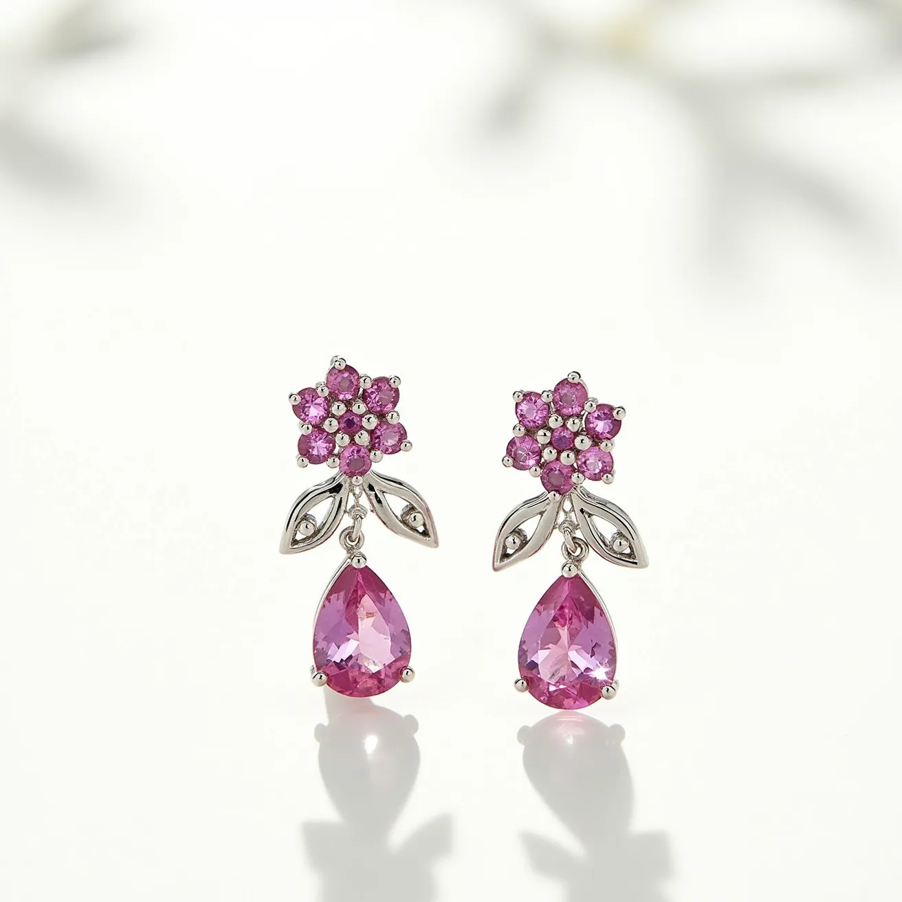 These pink sapphire earrings feature a charming design with floral and pear elements. The top part of the earrings is crafted in the shape of a flower, composed of multiple small round-cut pink sapphires clustered together, which create a vibrant, sparkling display. Beneath the floral arrangement are two small metallic leaves, likely made from a precious metal such as silver or white gold, adding a delicate touch to the overall design. The earrings are further enhanced by a larger, pear-shaped pink sapphire that dangles gracefully below, securely set in a three-prong setting. This combination of settings and cuts showcases the vibrant color and brilliance of the sapphires, making these earrings a striking accessory.