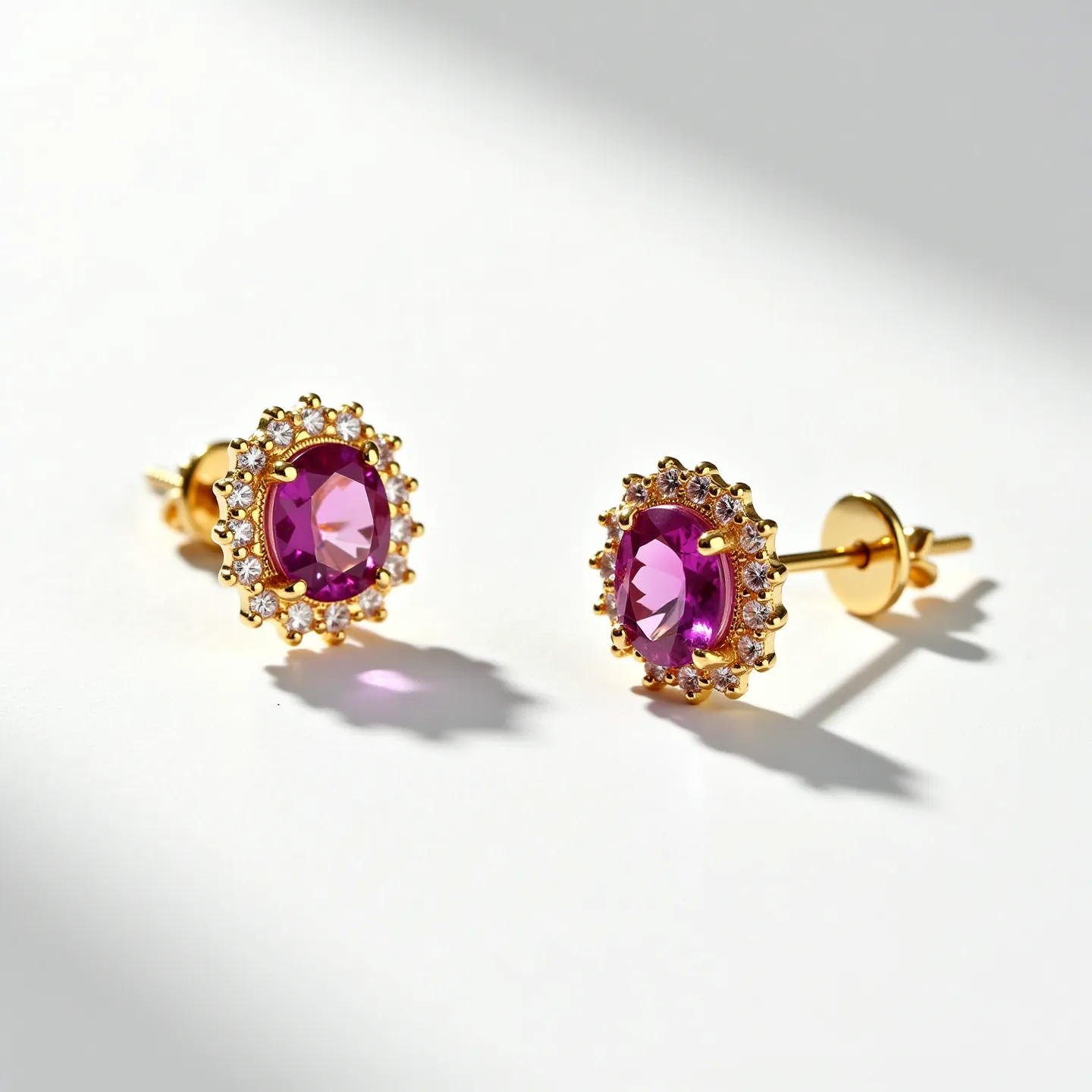 These pink sapphire earrings feature a central oval-cut pink sapphire, elegantly set in a surrounding halo of small round brilliant-cut diamonds. The gems are set in a polished yellow gold, enhancing the vividness of the stones. The earrings are of the stud type, secured with a post and butterfly clasp for a traditional and secure attachment. The intricate craftsmanship highlights the contrast between the vibrant pink center stone and the sparkling surrounding diamonds, creating a striking and classic design.