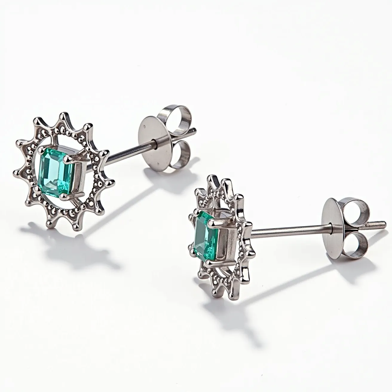 These post earrings feature a striking design with a central emerald-cut green stone that is securely set in a four-prong setting. The stone is encircled by a decorative metal frame with a sunburst pattern, adding a touch of elegance and complexity. The material of the earrings appears to be a reflective silver-tone metal, which complements the vibrant color of the green gemstone. The attachment mechanism is a standard push-back clasp, ensuring a secure and comfortable fit for the wearer. The combination of the unique geometric pattern and the bright green centerpiece creates a sophisticated and appealing aesthetic.