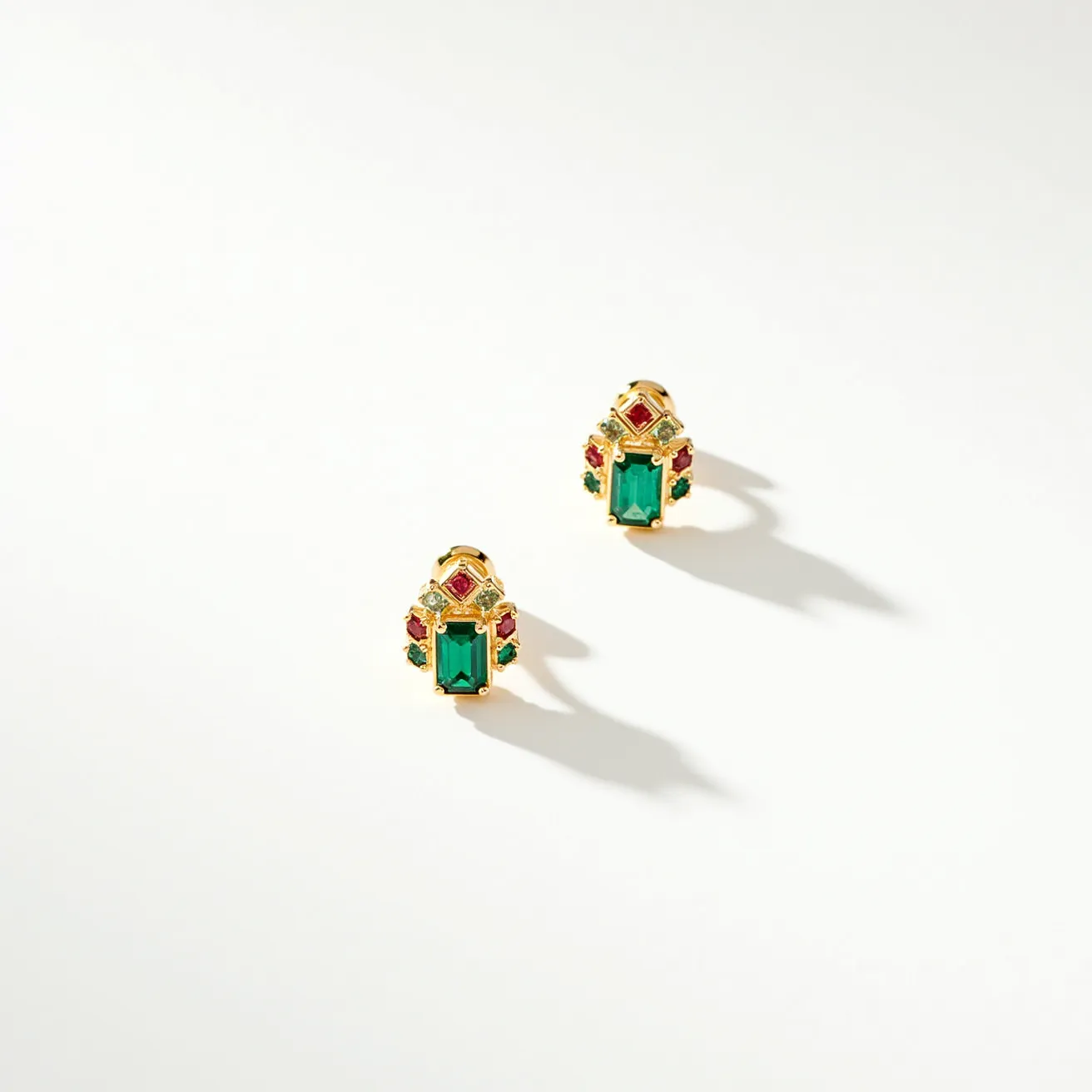 These post earrings are intricately designed with a central emerald-cut green stone set in a bezel setting, creating a prominent focal point. Surrounding the green stone are smaller round and square stones in red and green hues, complementing the central stone and adding a vibrant contrast. The settings are crafted from a gold-toned metal, providing a rich, warm backdrop to the colorful stones. These earrings attach with a traditional post, ensuring secure placement when worn. The combination of colorful stones and gold-toned metal gives these earrings a regal and elegant appearance.