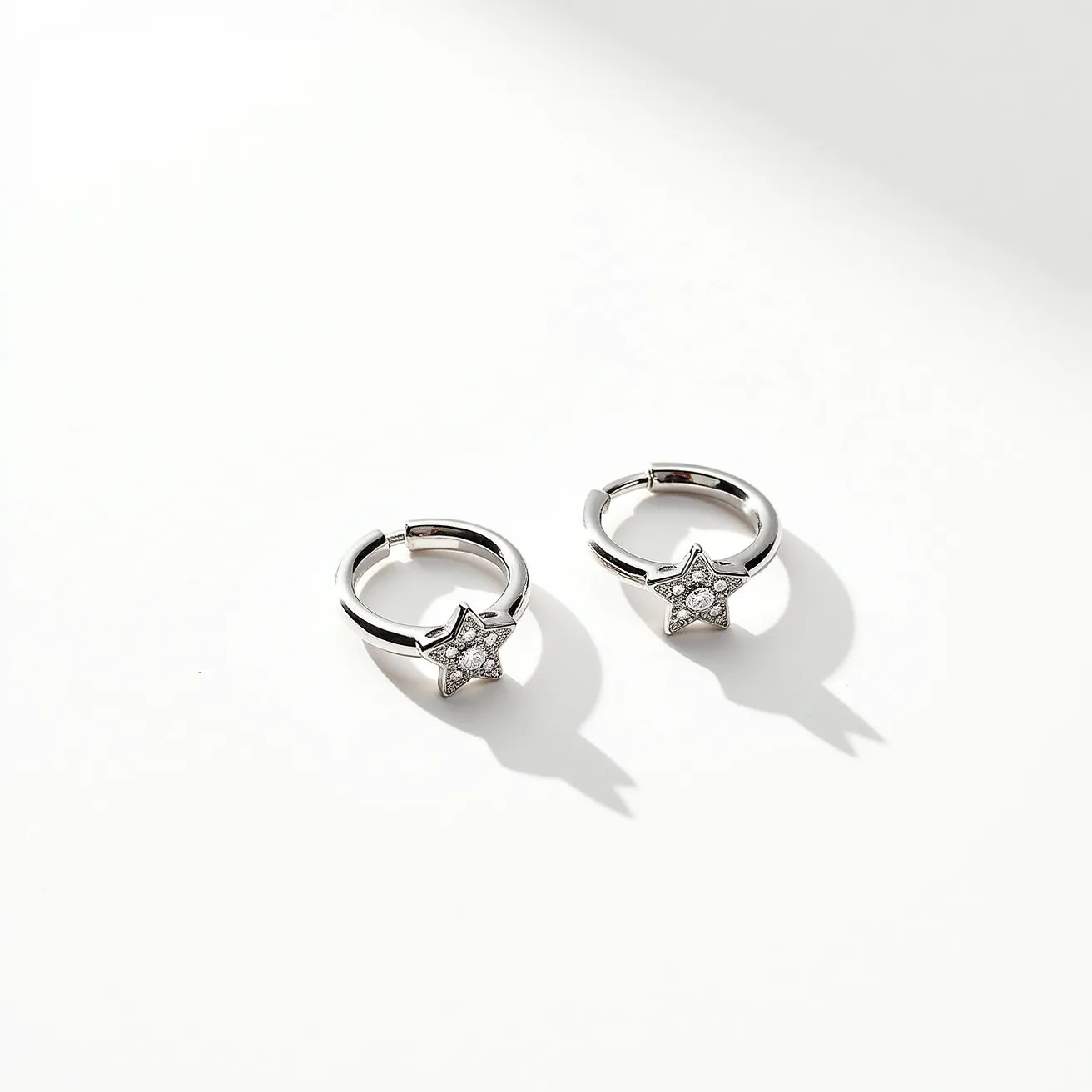 These ring earrings feature a sleek metallic finish, likely composed of silver or white gold, forming a classic hoop design. Each earring is adorned with a star-shaped charm at the base, embellished with several small, clear gemstones, possibly diamonds or cubic zirconia, set in a pavé arrangement to enhance sparkle and brilliance. The closure mechanism appears to be a hinge clasp, providing secure attachment while maintaining ease of use. The overall design offers a contemporary yet elegant aesthetic, suitable for both everyday wear and special occasions.