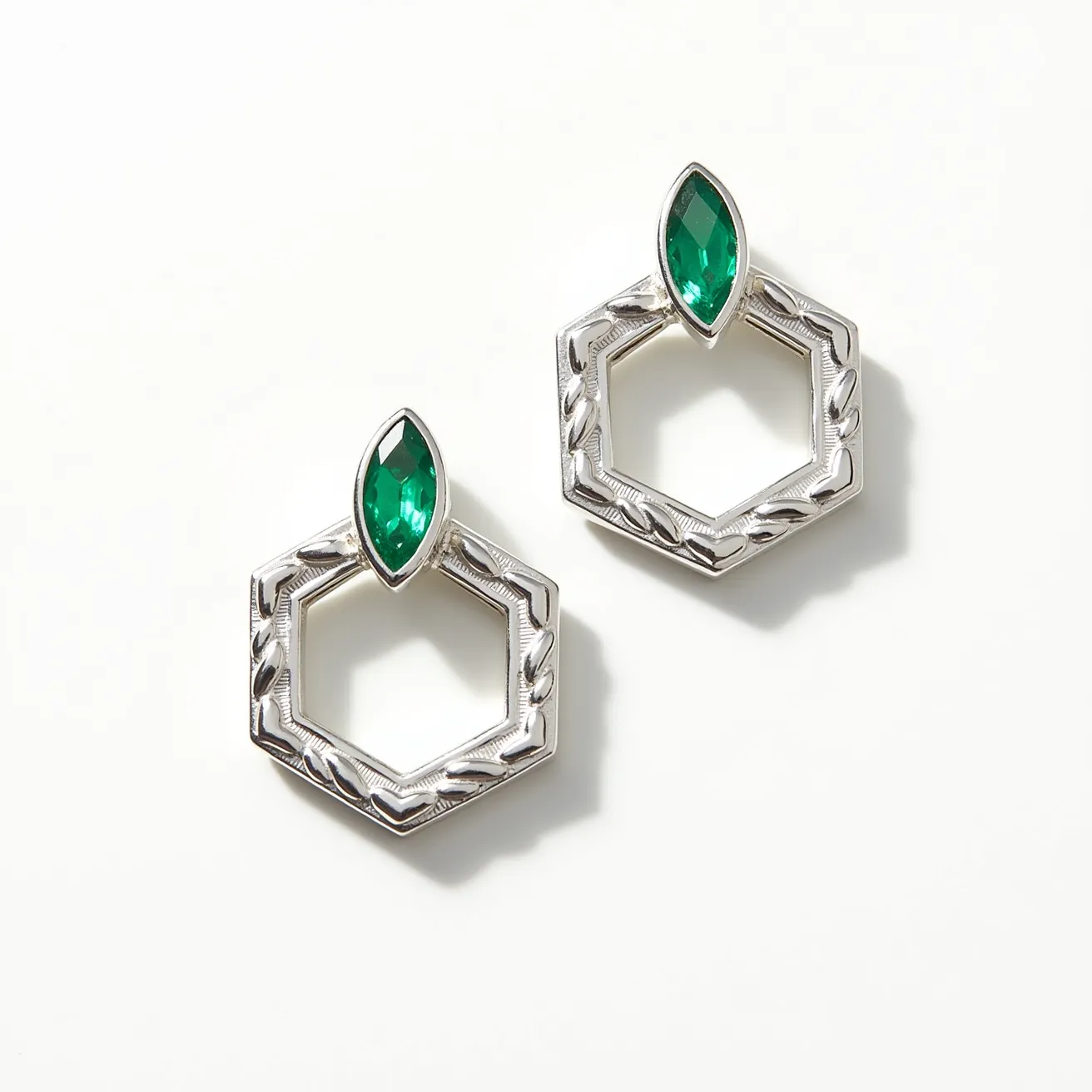 These ring earrings feature an elegant hexagonal hoop design crafted from what appears to be a polished metal, possibly silver, exhibiting a textured pattern along the edges. Each earring is adorned with a marquise-cut green gemstone, possibly an emerald or similar stone, set in a bezel setting at the top vertex of the hexagon. The gemstone's marquise cut maximizes brilliance and adds a sophisticated touch to the design. The earrings are likely secured with a post or stud, although the exact clasp or attachment isn't clearly visible. The refined combination of green stone and metallic texture provides a striking and modern aesthetic.