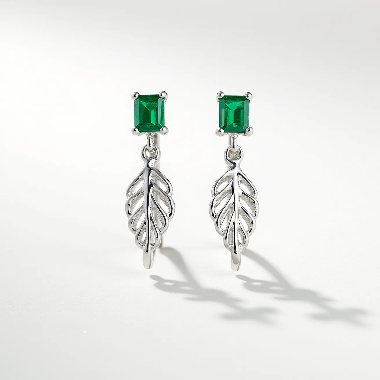 These ring earrings feature a striking design with each piece showcasing a square-cut green gemstone, likely emerald, set in a four-prong silver setting. The earrings have a drop-style attachment, which includes a delicately crafted silver leaf motif hanging elegantly beneath each gem. The combination of the vivid green stones and the intricate silver leaves creates a beautiful and elegant look. The attachment appears to be a hook or similar type, allowing the earrings to dangle gracefully when worn.