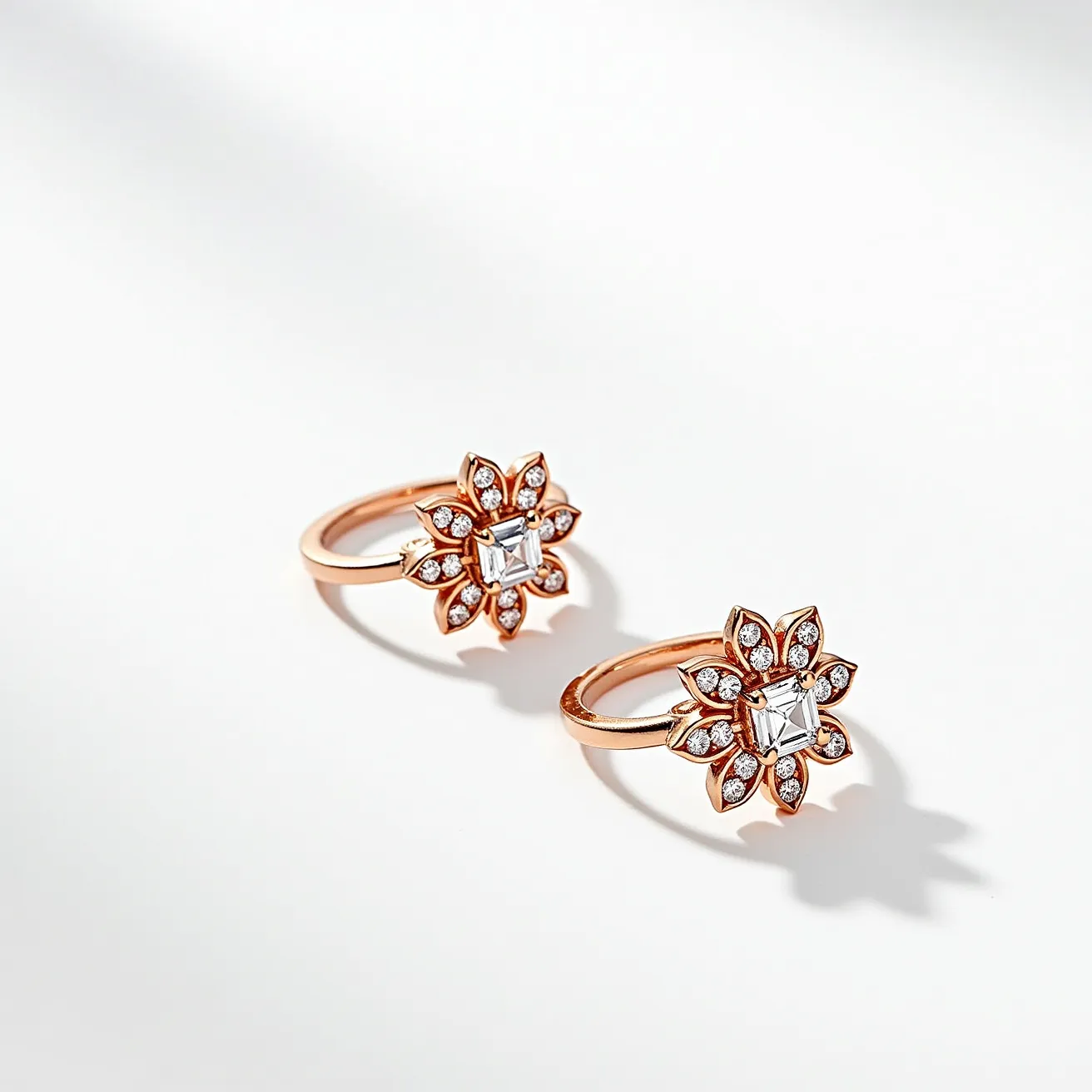 These ring earrings feature an elegant floral design crafted from a warm, rose gold material. Each earring showcases a central emerald-cut gem, likely a clear diamond or similar stone, set in a prong setting that emphasizes its clarity and brilliance. Surrounding the central stone are multiple smaller round-cut diamonds, each securely nestled in petal-inspired settings that add a delicate sparkle. The hoops are smoothly polished, enhancing the overall luxurious appearance. The earrings have a secure latch back clasp, ensuring they are both stylish and functional.