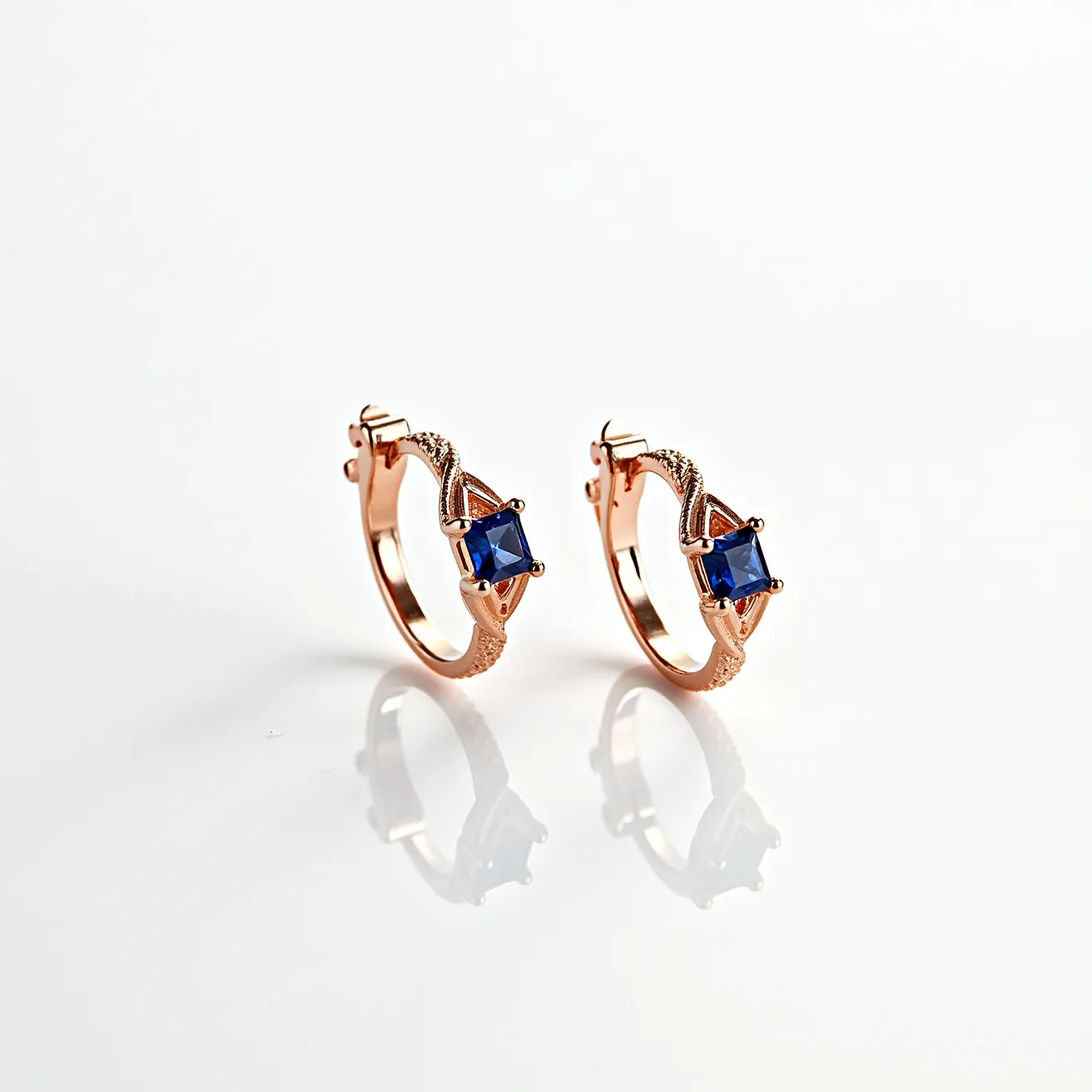 These ring earrings are crafted from a rose gold metal, featuring a central square-cut blue gemstone, likely a blue sapphire, set in a prominent prong setting that enhances the gem's visibility and elegance. The rose gold complements the deep blue of the stones, adding a warm contrast to the overall design. The earrings are designed with a hoop style that includes a secure hinged clasp, ensuring they remain comfortably fastened when worn. The intricate detailing on the metalwork surrounding the gemstone adds a unique touch, highlighting the earring’s luxurious appeal.