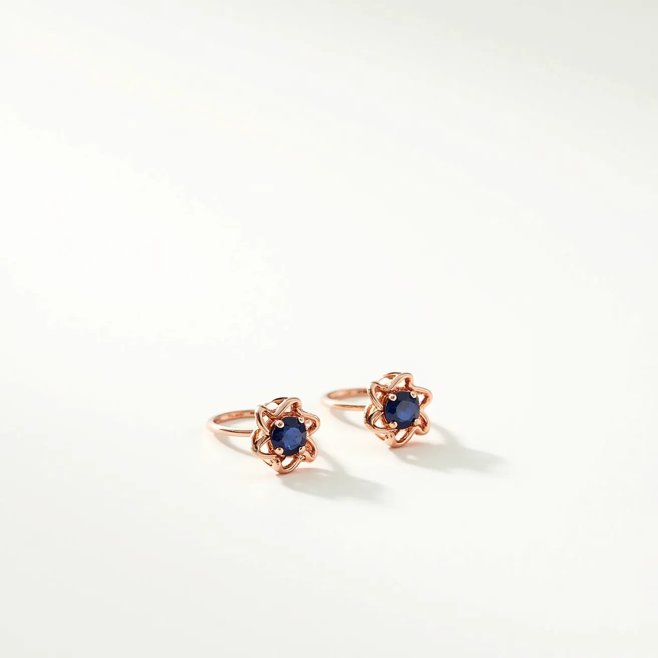 These ring earrings feature a striking design crafted from what appears to be a warm rose gold or similar metal, forming a delicate band. At their center, each earring holds a round, deep blue gemstone, likely a sapphire, set in a star-like prong setting that secures the stone while adding an ornate touch. The prongs enhance the overall elegance, creating a captivating interplay between metal and gem. The earrings are equipped with a simple hoop mechanism that allows for easy insertion and secure closure in pierced ears. The harmonious combination of the vivid blue gems and the lustrous metal results in a sophisticated and timeless accessory.