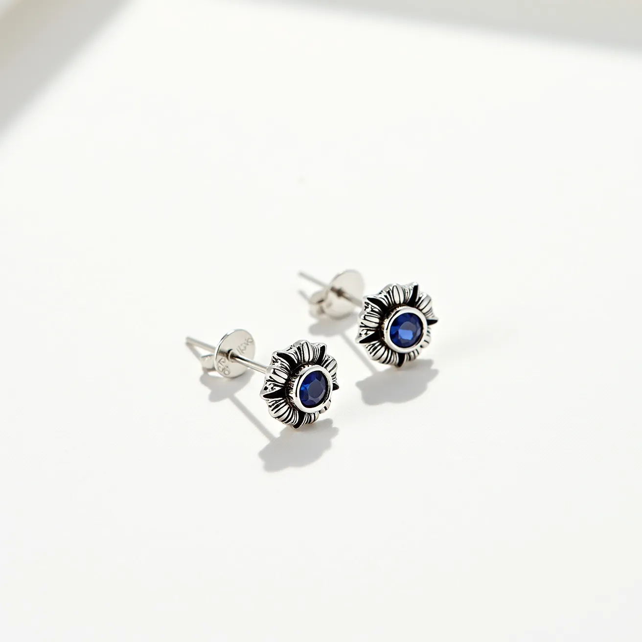 These rose earrings feature a striking floral design crafted from silver metal, exhibiting a detailed petal motif surrounding a central blue gemstone. The gemstone is round cut and prong set, adding a vibrant pop of color and sparkle to the earrings. The attachment is a classic post and butterfly back, ensuring a secure and comfortable fit for the wearer. These earrings beautifully combine elegance with an artistic floral touch, making them a versatile accessory for a variety of occasions.