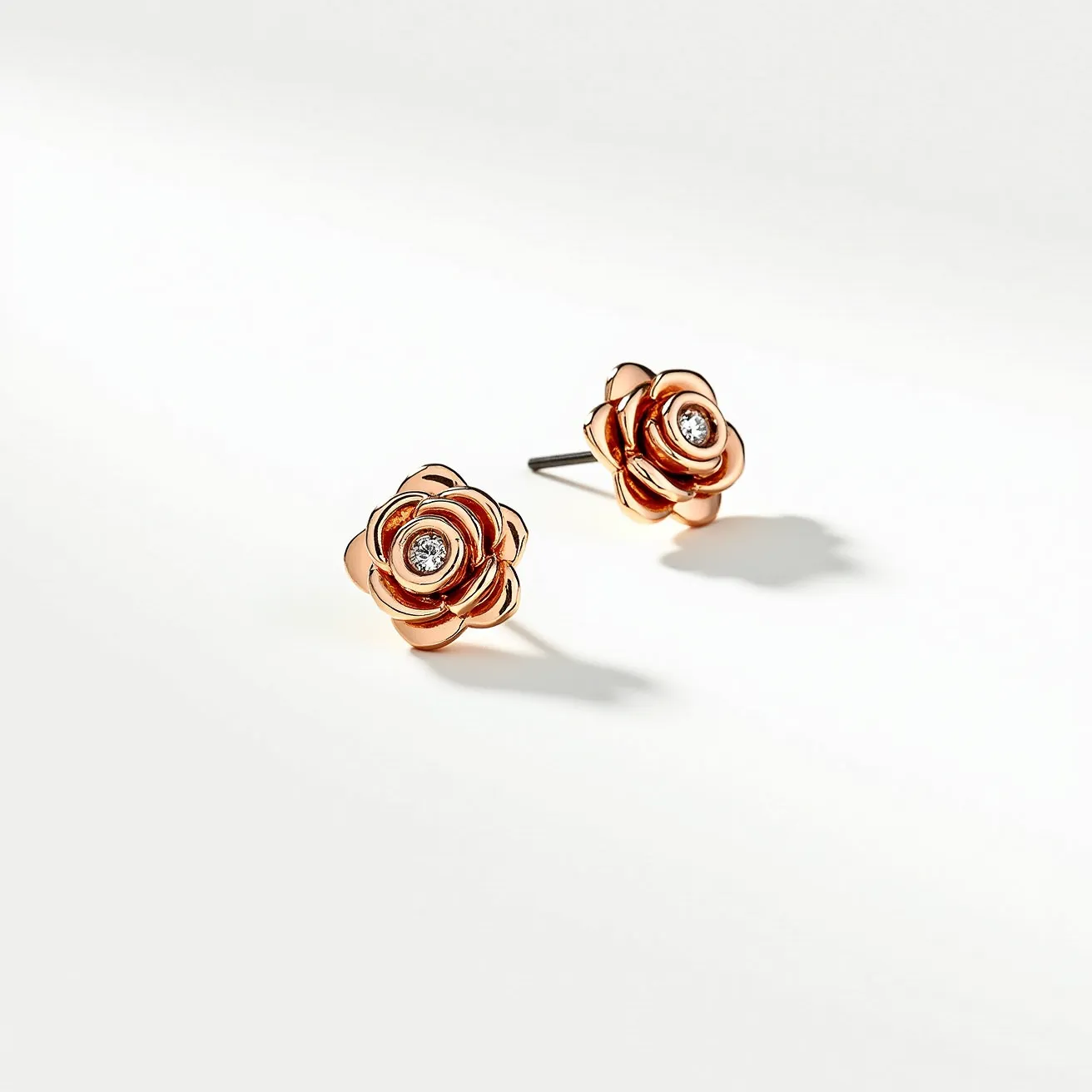 These rose earrings exhibit a charming floral design crafted from a polished rose gold material, giving them a luxurious and elegant appearance. Each earring features a central round-cut diamond set securely within the layers of the rose petals, adding a touch of sparkle and enhancing their sophisticated allure. The earrings are designed with a simple stud post, allowing them to be worn comfortably with a secure friction back clasp.